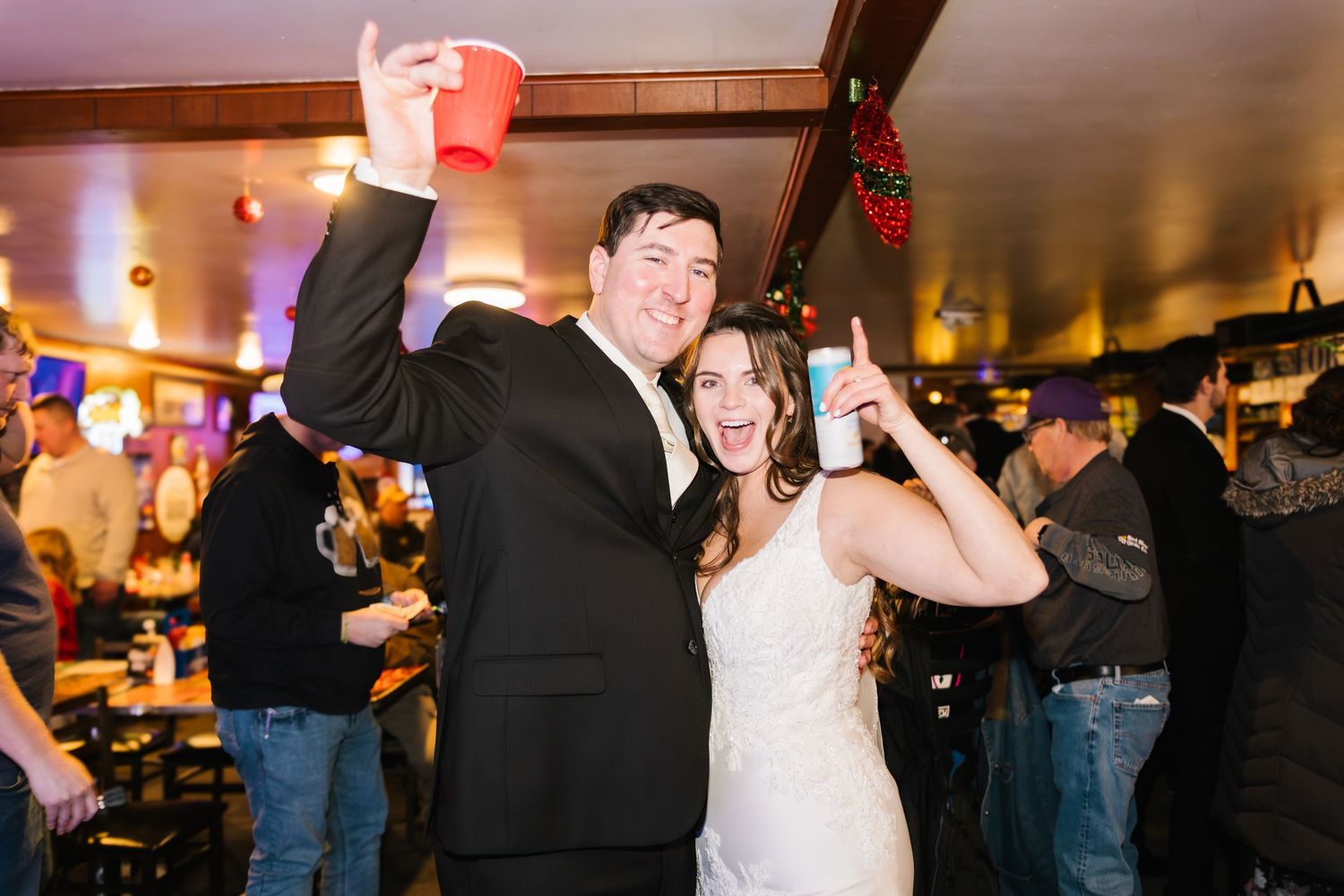Bride and groom celebrating at wedding reception, raising drinks and smiling in a festive bar venue.