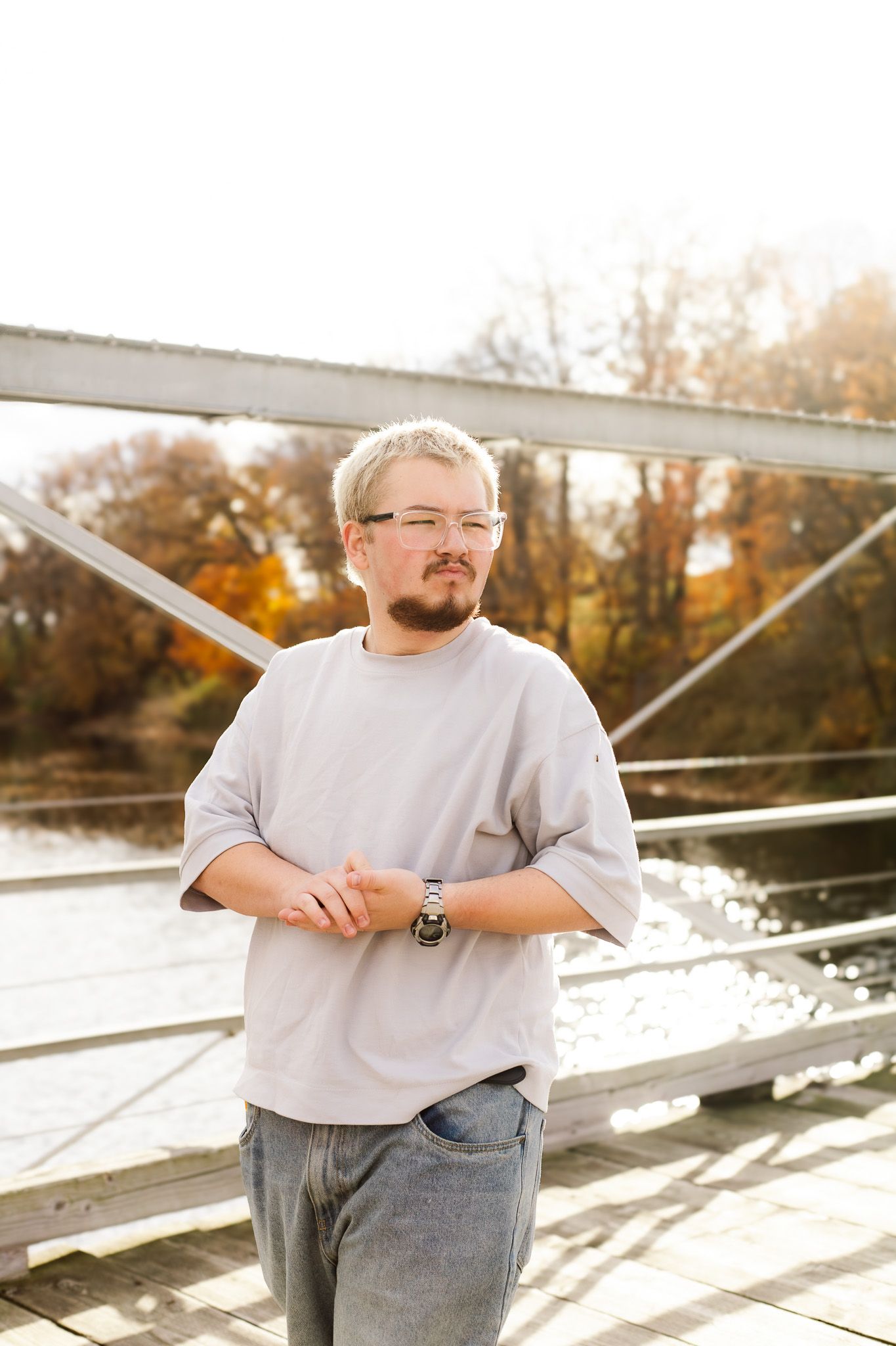 Man with glasses and beard in gray t-shirt standing on a bridge with autumn trees and river in background.