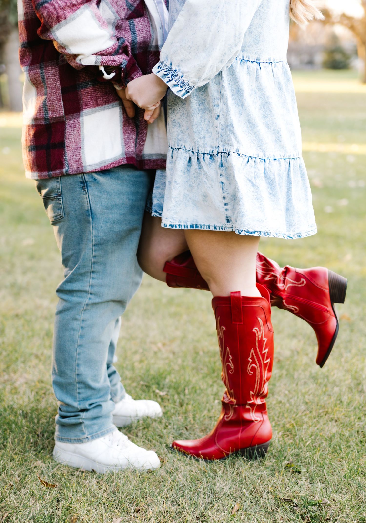 Couple holding hands outdoors, woman wearing red cowboy boots and denim dress, man in plaid shirt and jeans on green grass.