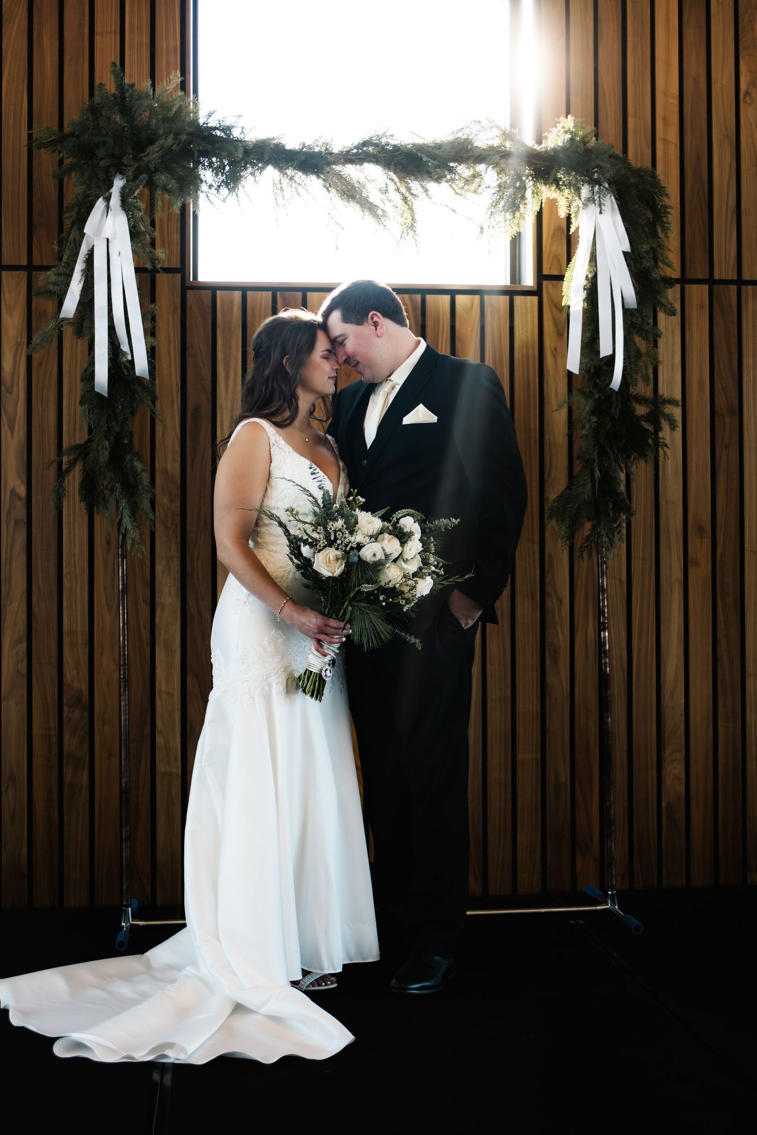 Bride and groom share romantic moment at rustic wooden altar adorned with greenery and white ribbon bows.