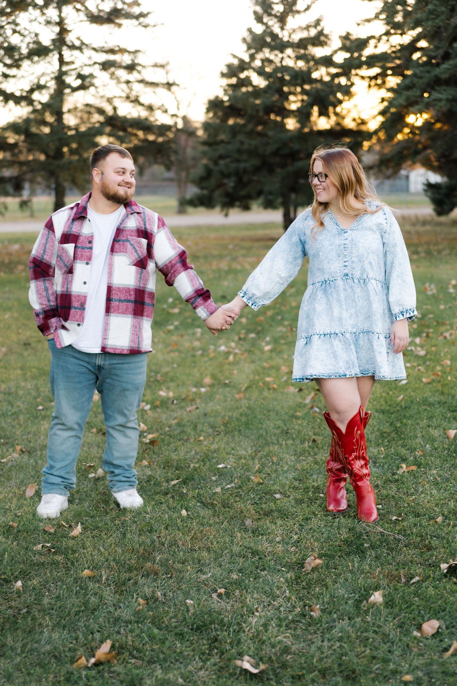 Couple holding hands in a park at sunset, man in plaid shirt and jeans, woman in denim dress and red cowboy boots.