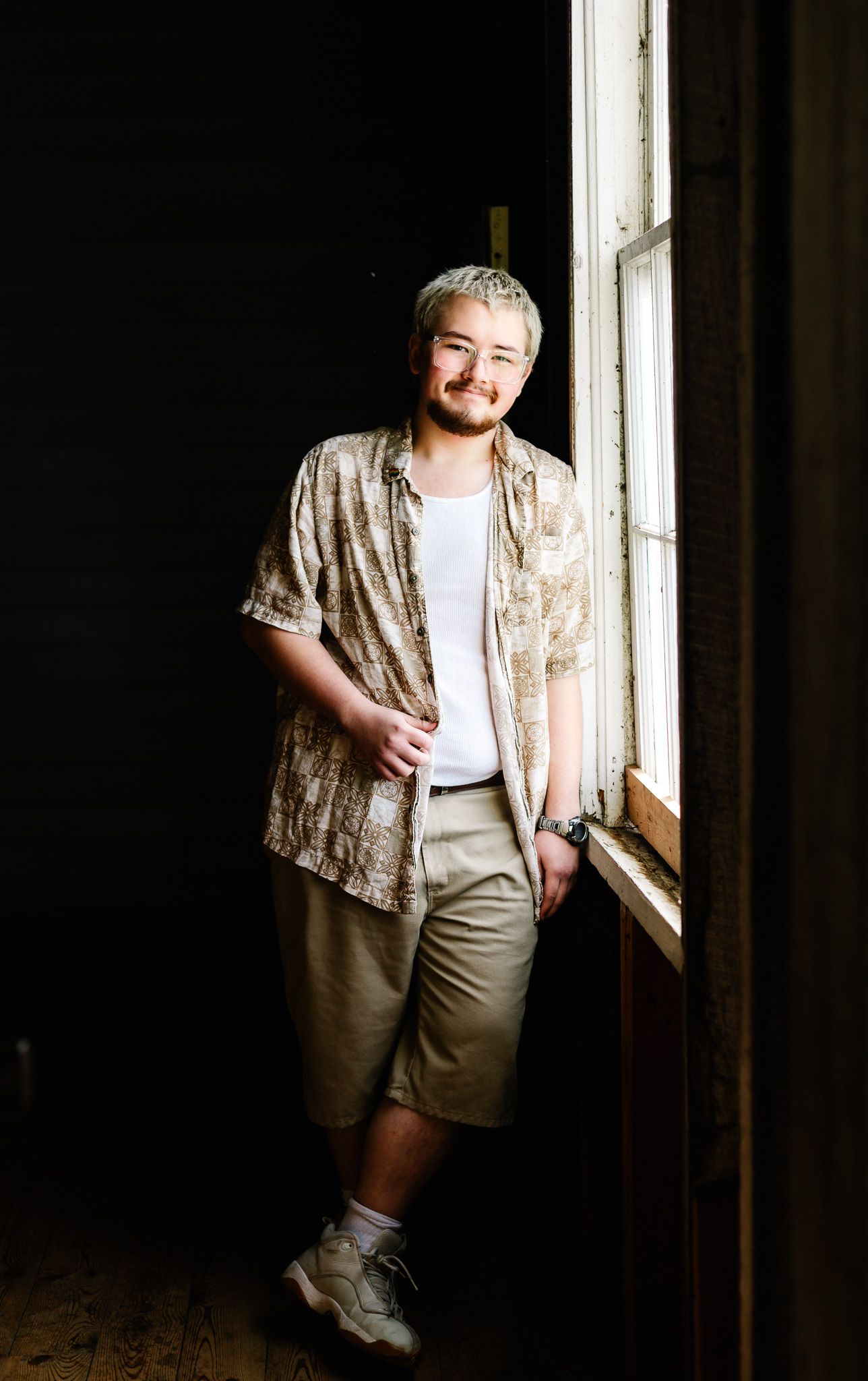 Young man with blonde hair smiling by a window, wearing a patterned shirt, white tee, khaki shorts, and boots.