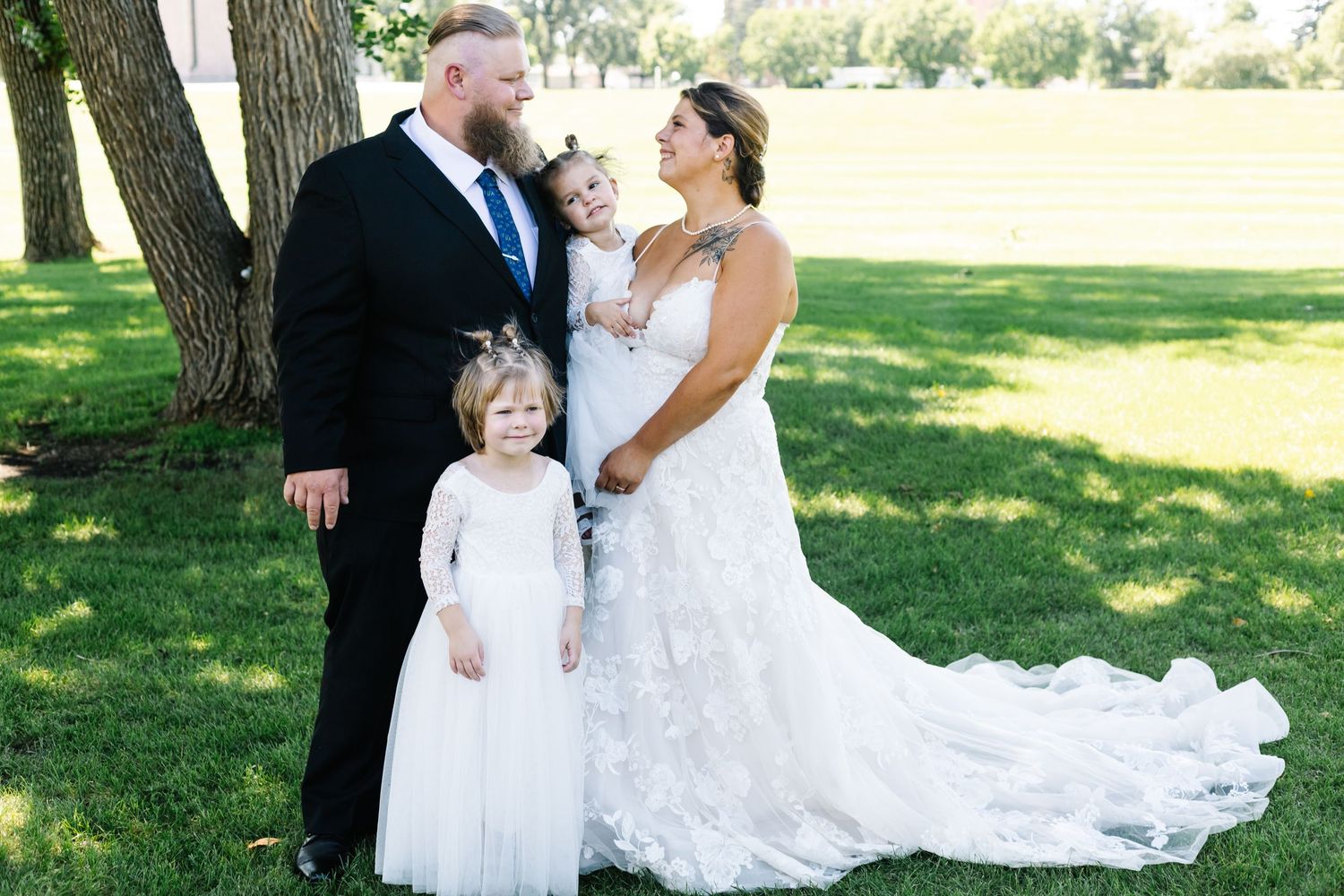 Wedding party poses outdoors with bride in flowing white gown under shady trees.