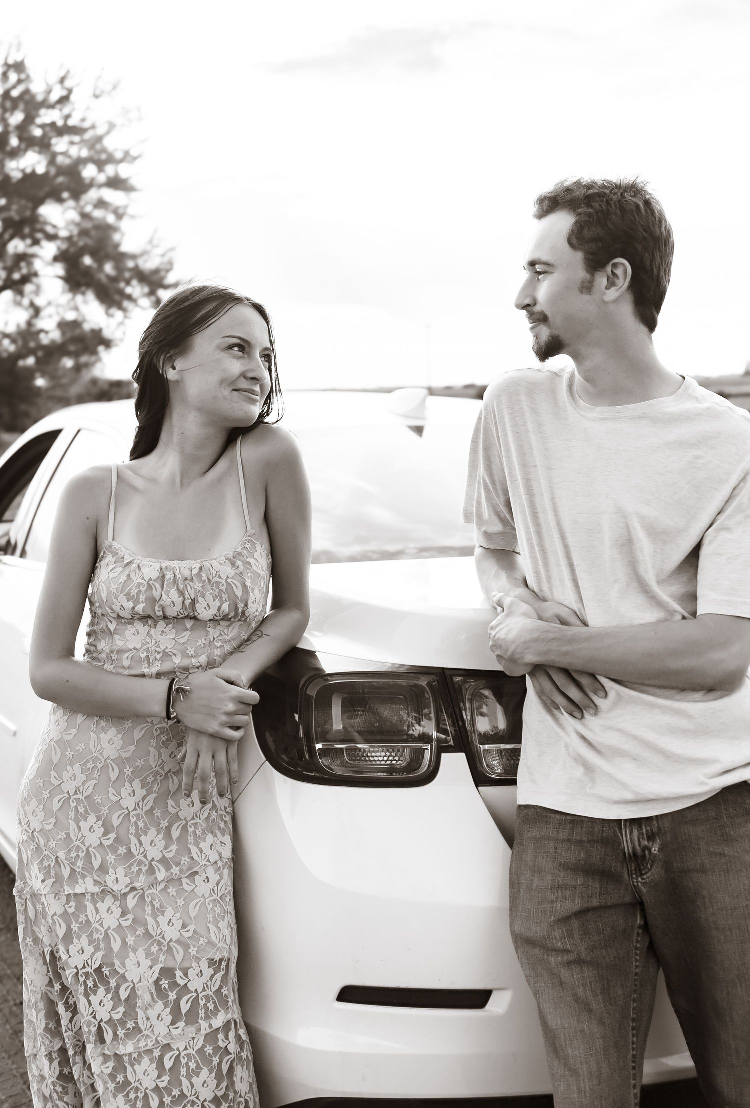 A couple shares candid moments by their parked car in black and white.