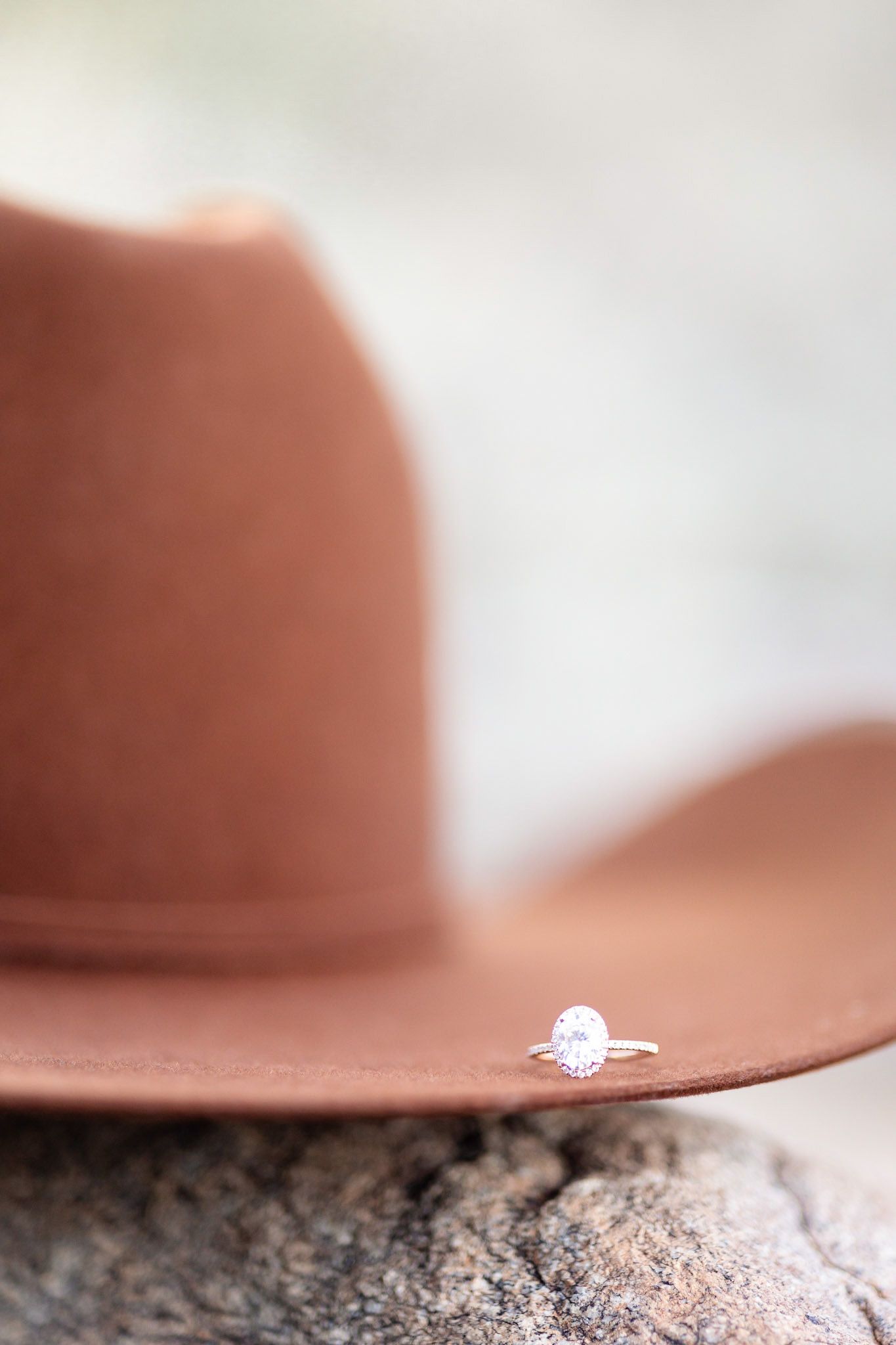 A sparkling diamond engagement ring rests delicately on the brim of a brown cowboy hat.