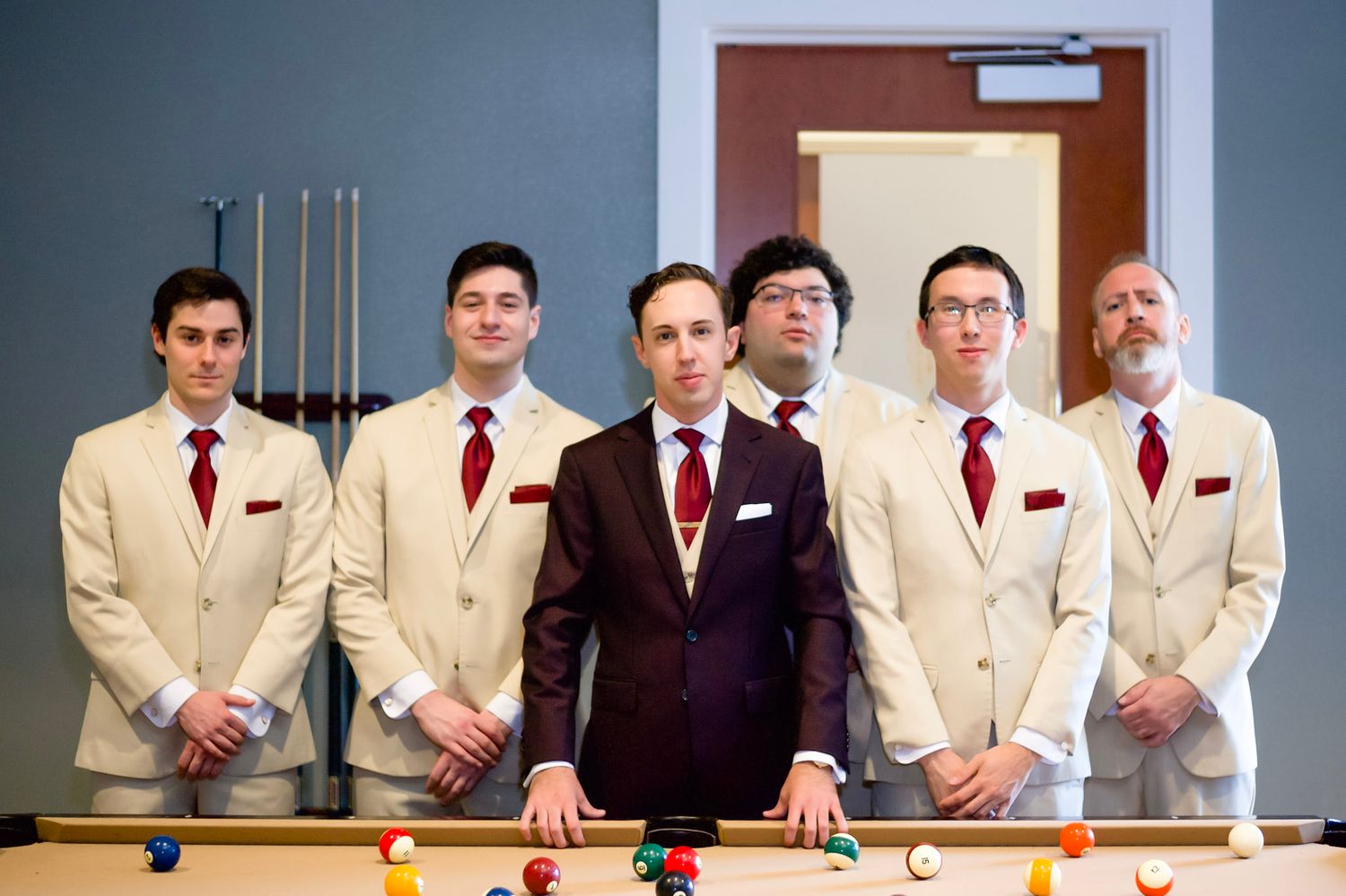 Groomsmen lined up wearing cream colored suits with burgundy ties at a formal wedding.