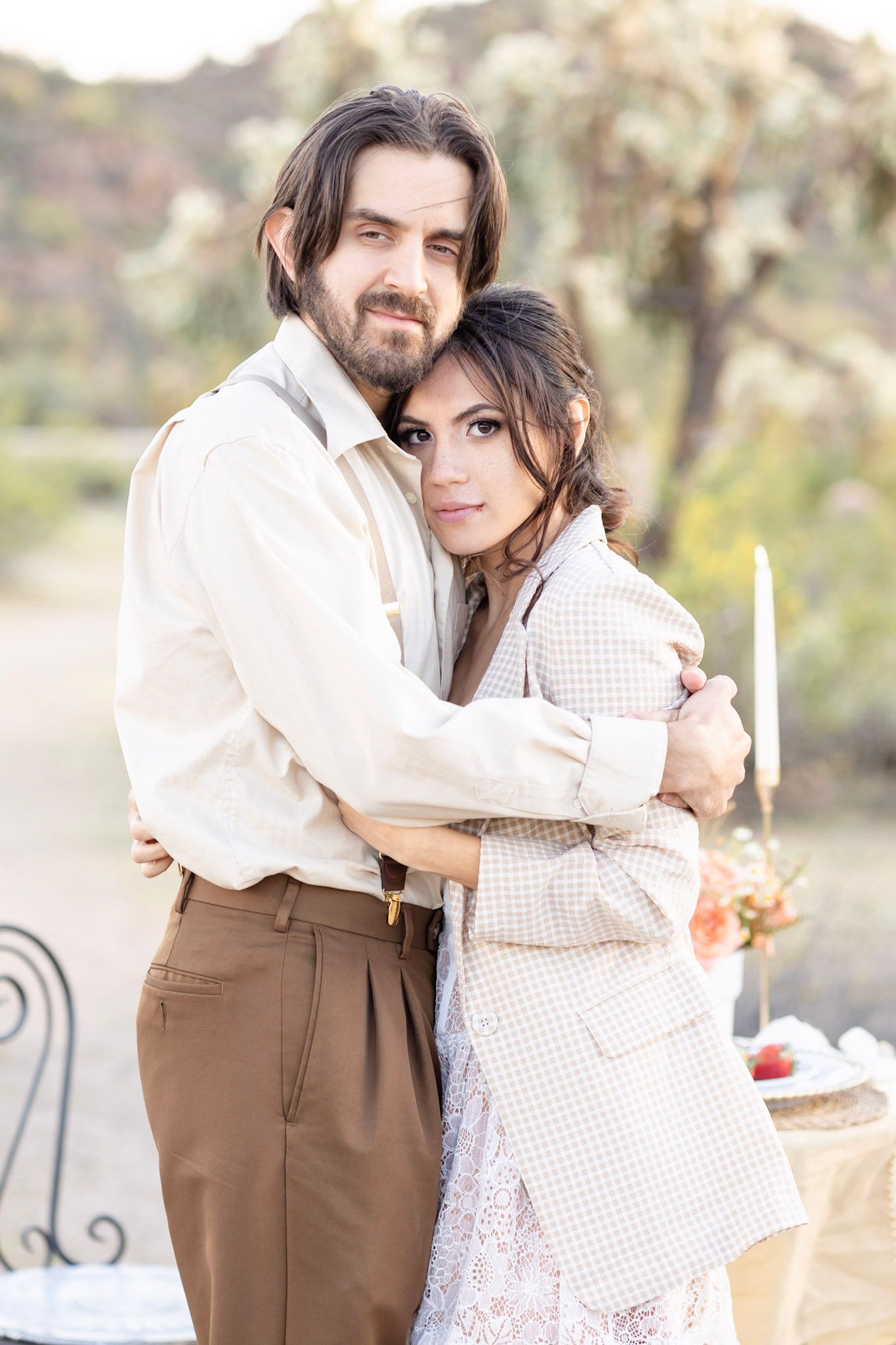 Romantic vintage-inspired couple embraces in a rustic outdoor setting wearing white and brown attire.