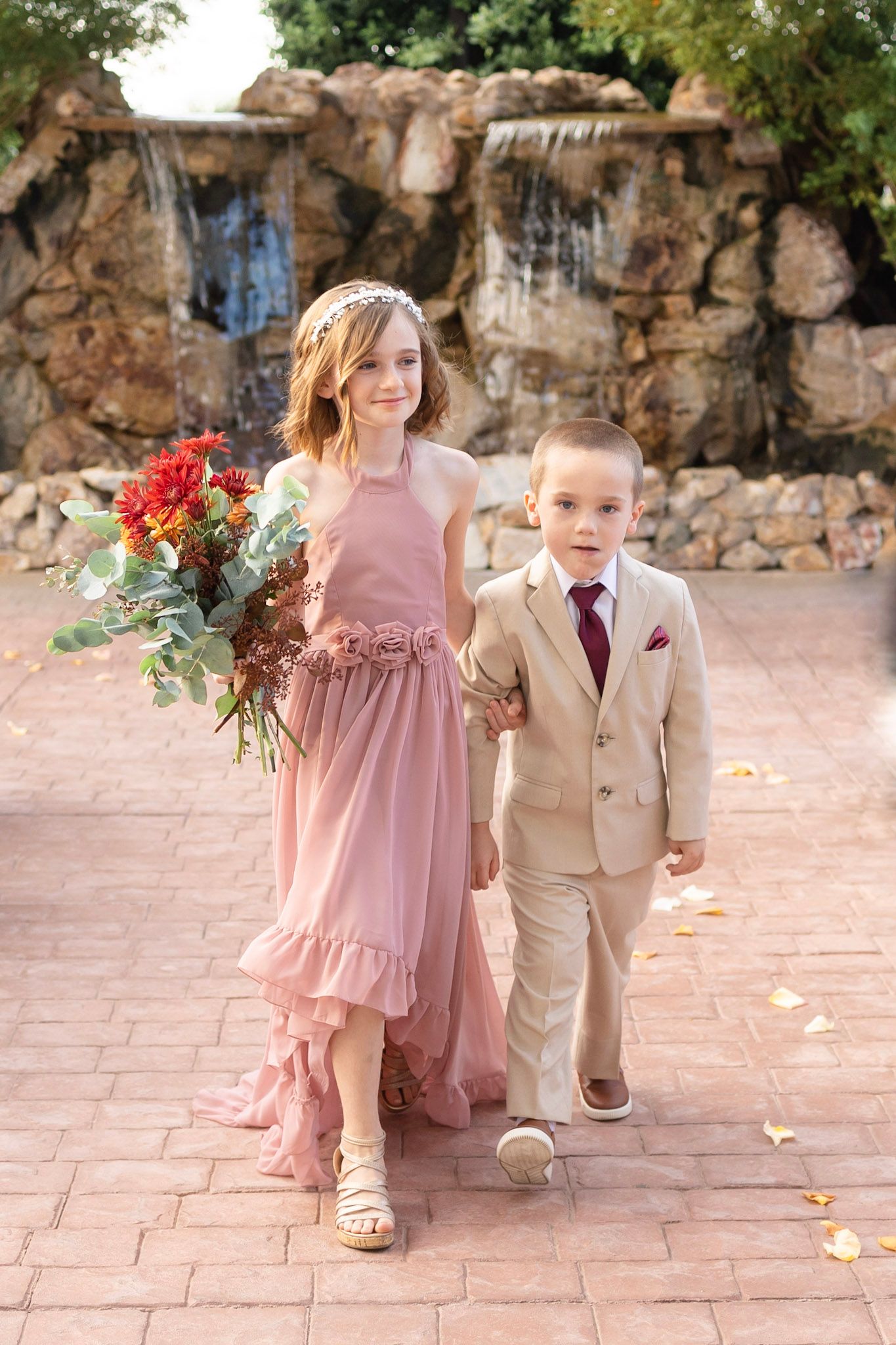 Young attendants in pink dresses and tan suits walk down a stone pathway at an outdoor wedding.