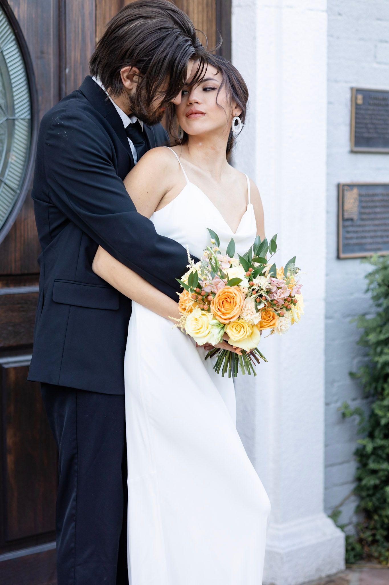 Wedding couple embraces near white building with peach and yellow bouquet.