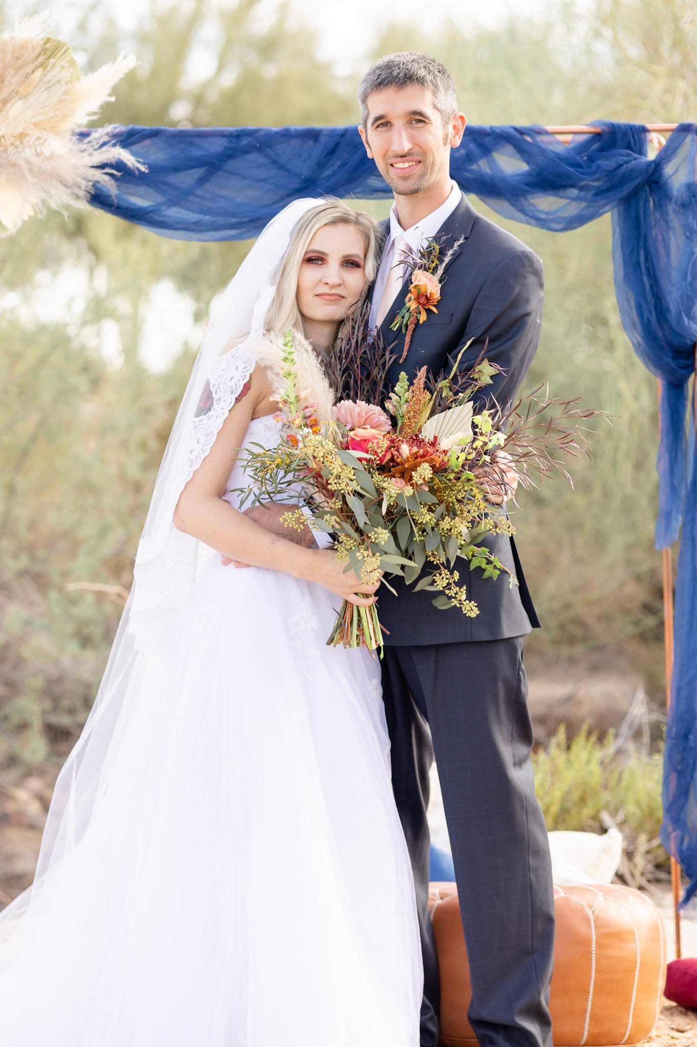 Wedding couple poses with fall-themed bouquet under blue fabric ceremony backdrop.
