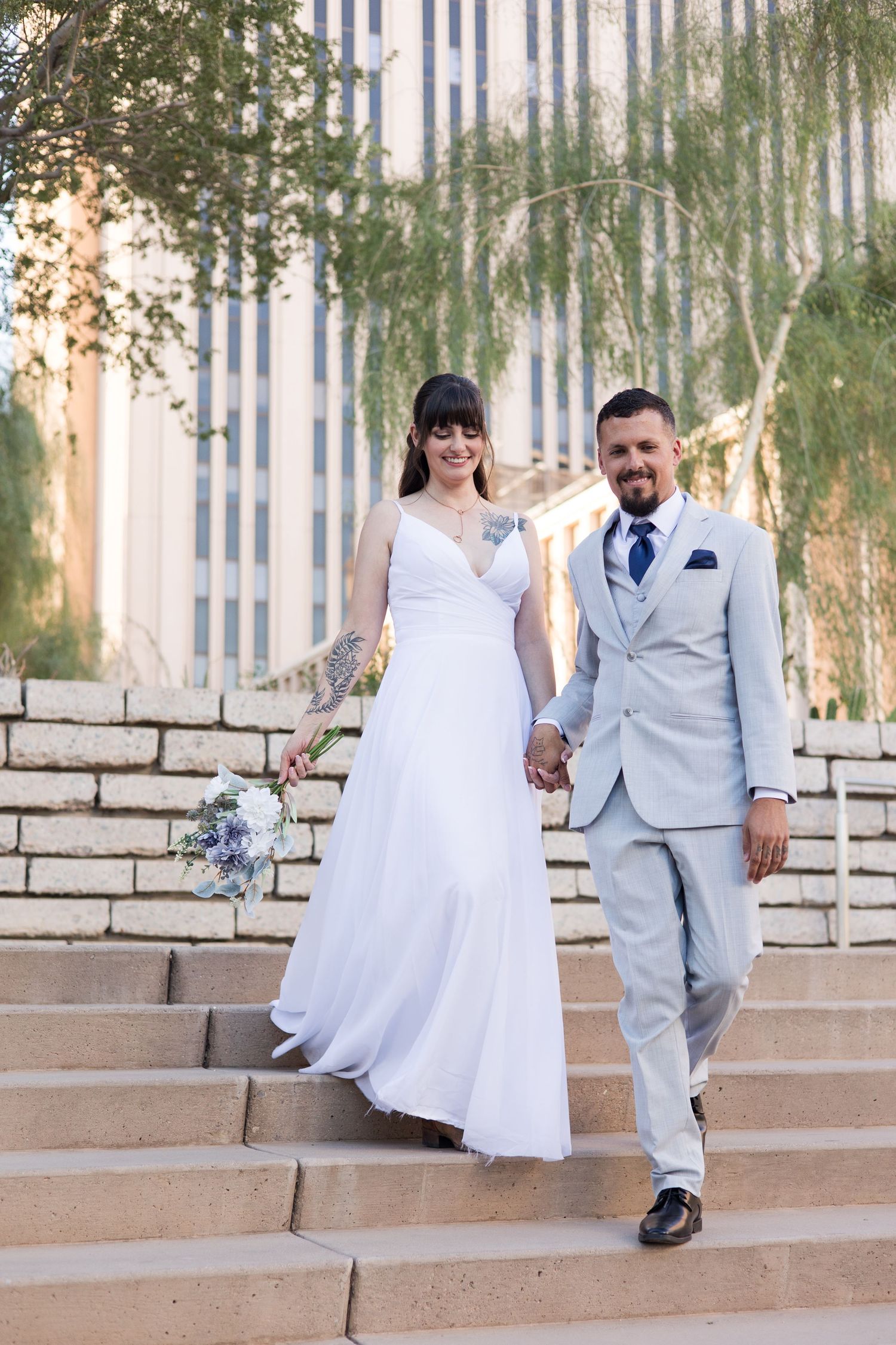 Couple in wedding attire sits casually on stone steps with trees in background.
