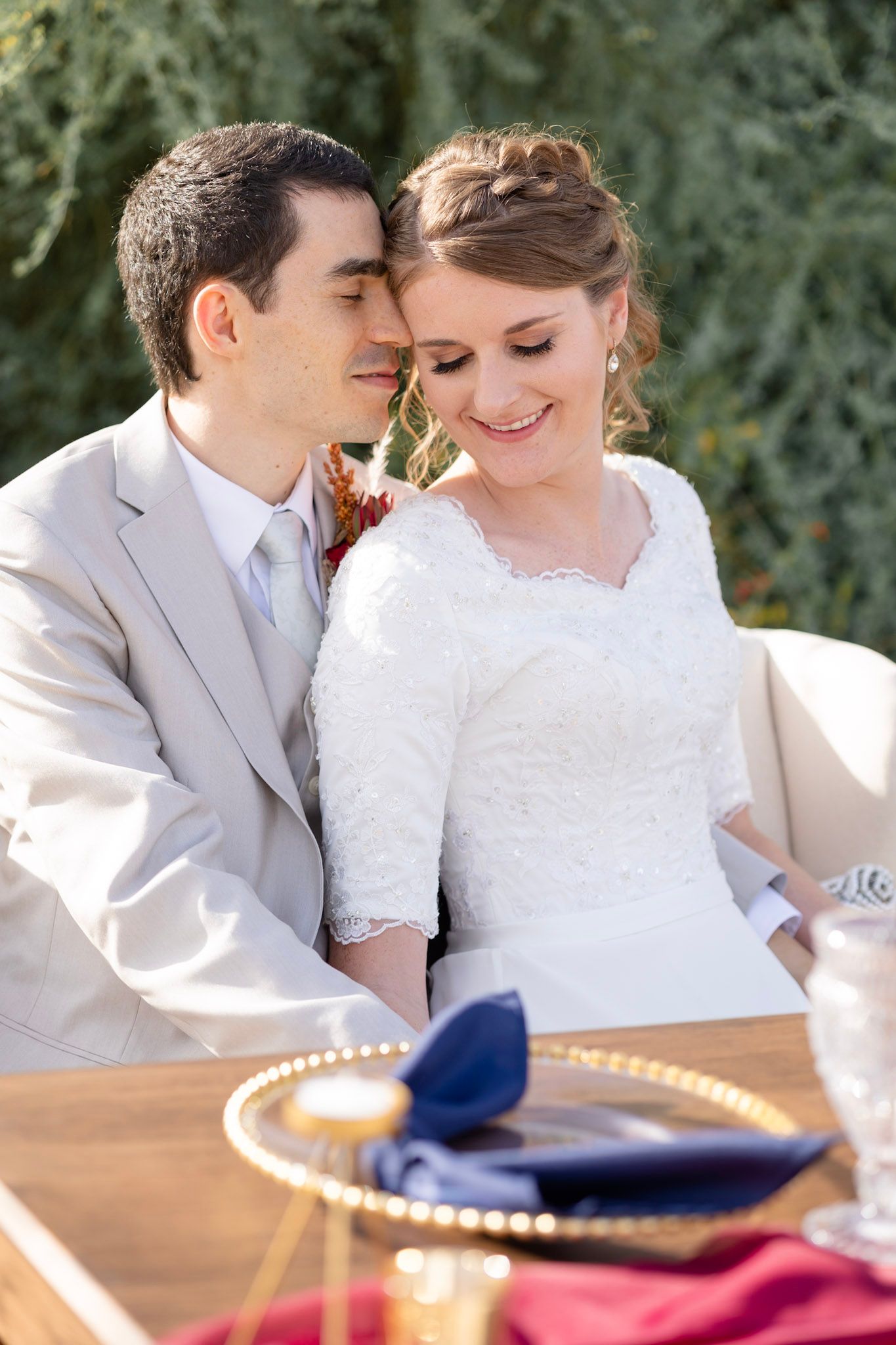 Newlyweds share tender moments at outdoor reception table with navy and red place settings.
