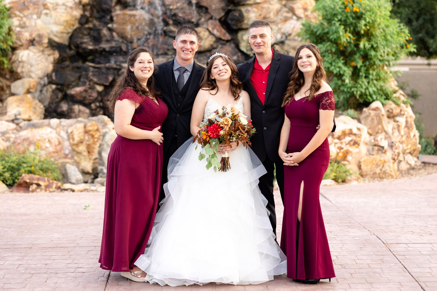 Wedding party wearing burgundy dresses and dark suits pose together outdoors.