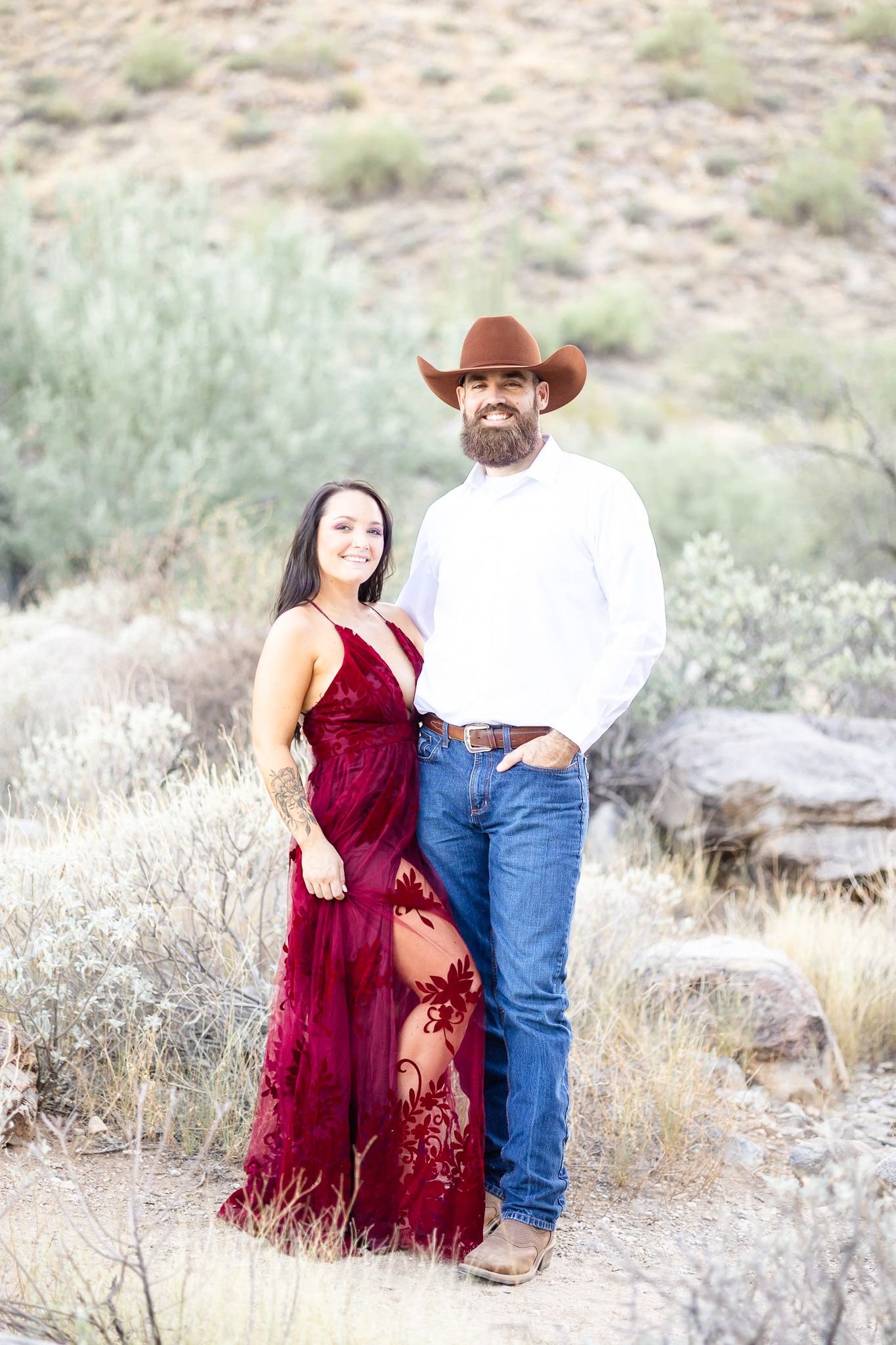 A couple in western attire poses together in a desert landscape with sagebrush and rocks.