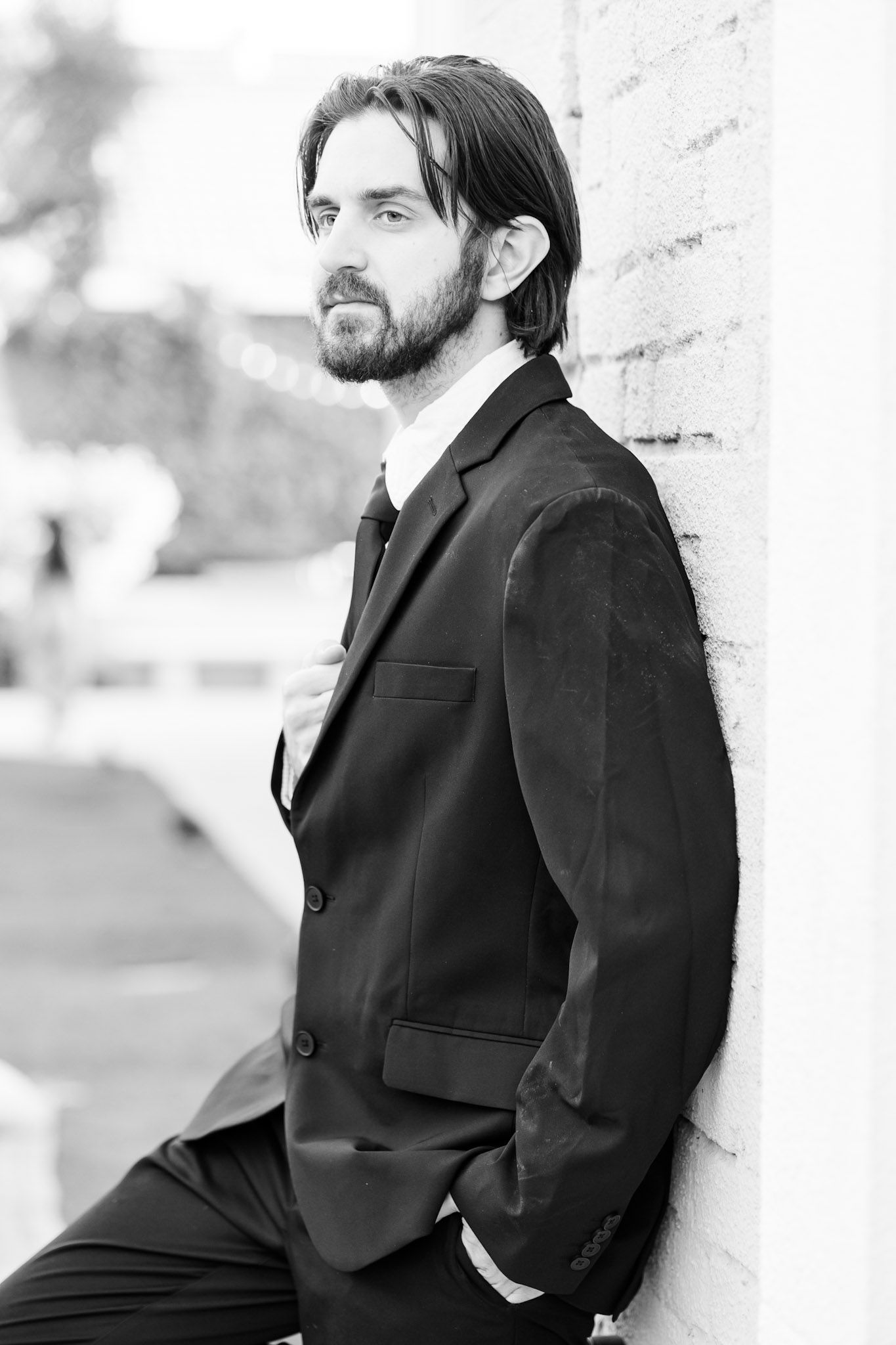 Black and white portraits of groom in suit leaning against white wall.