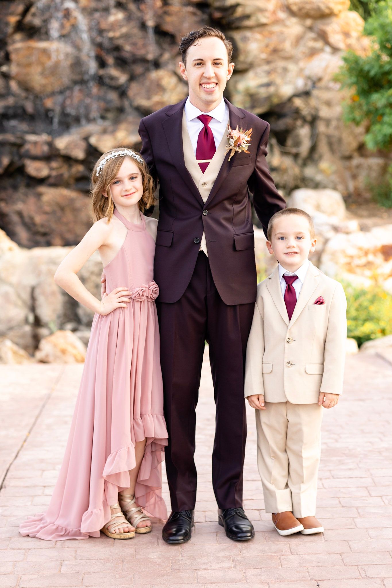 Wedding party members in pink dresses and dark suits pose together against a stone wall backdrop.