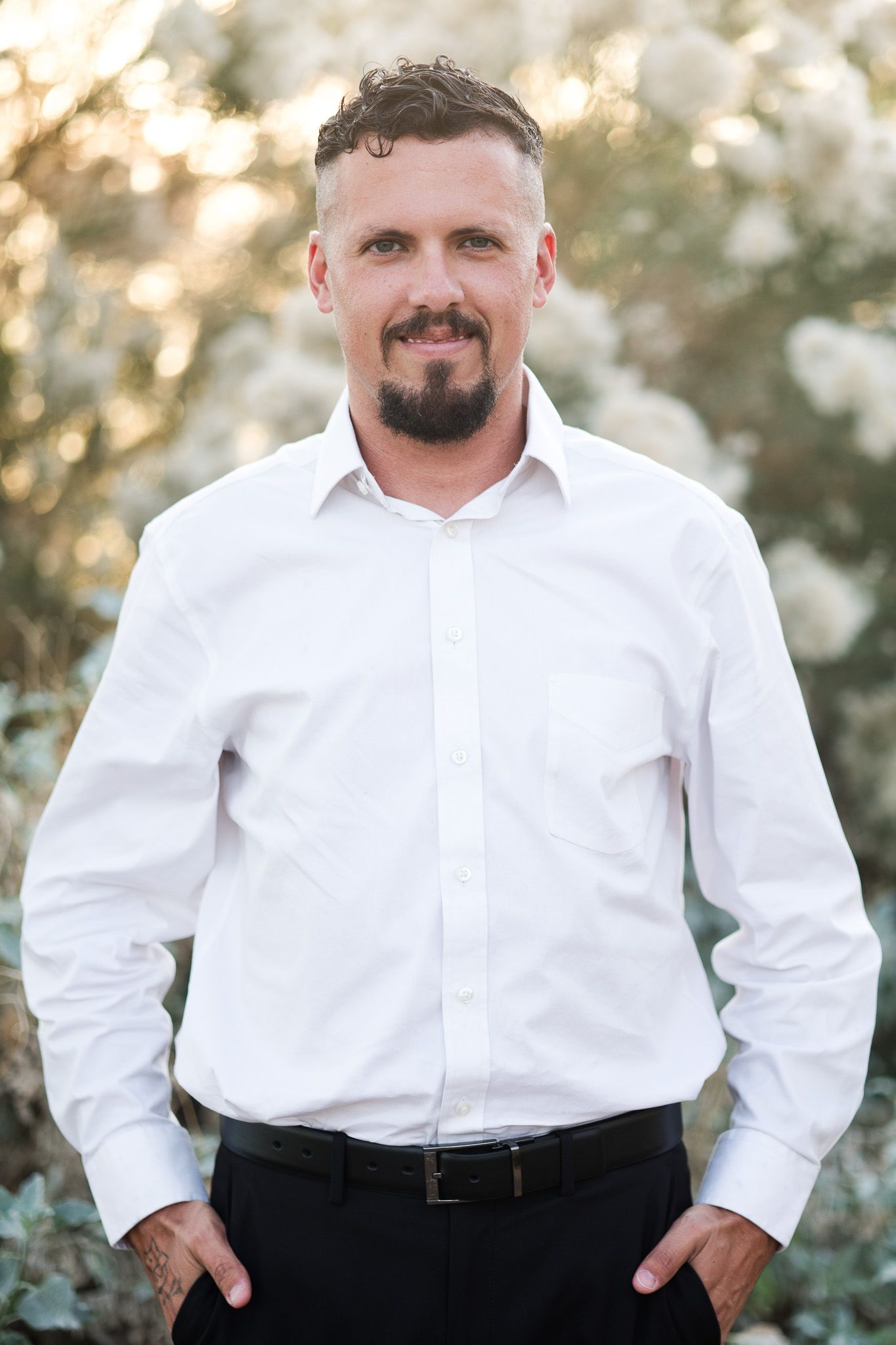 Professional portrait of someone in a crisp white dress shirt and dark pants against a soft bokeh background outdoors.