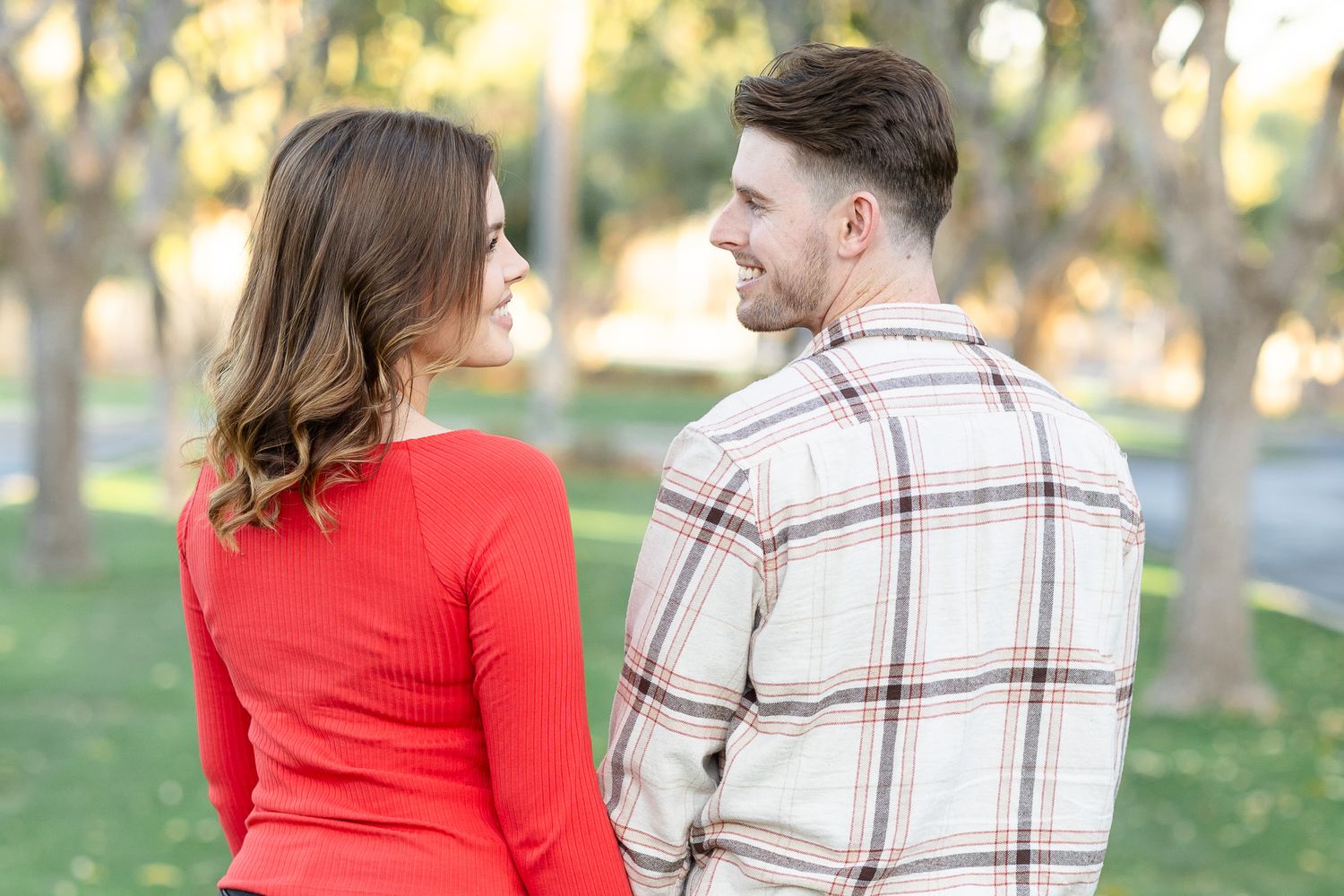 A couple in a plaid shirt and red sweater share a romantic moment together in a sunlit park.
