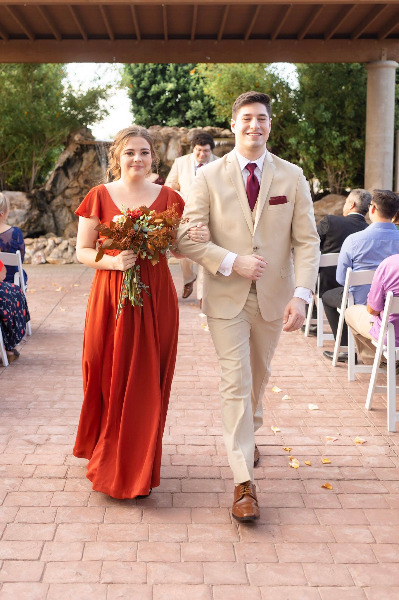 Wedding party wearing red dresses and beige suits walking down a stone courtyard during ceremony.