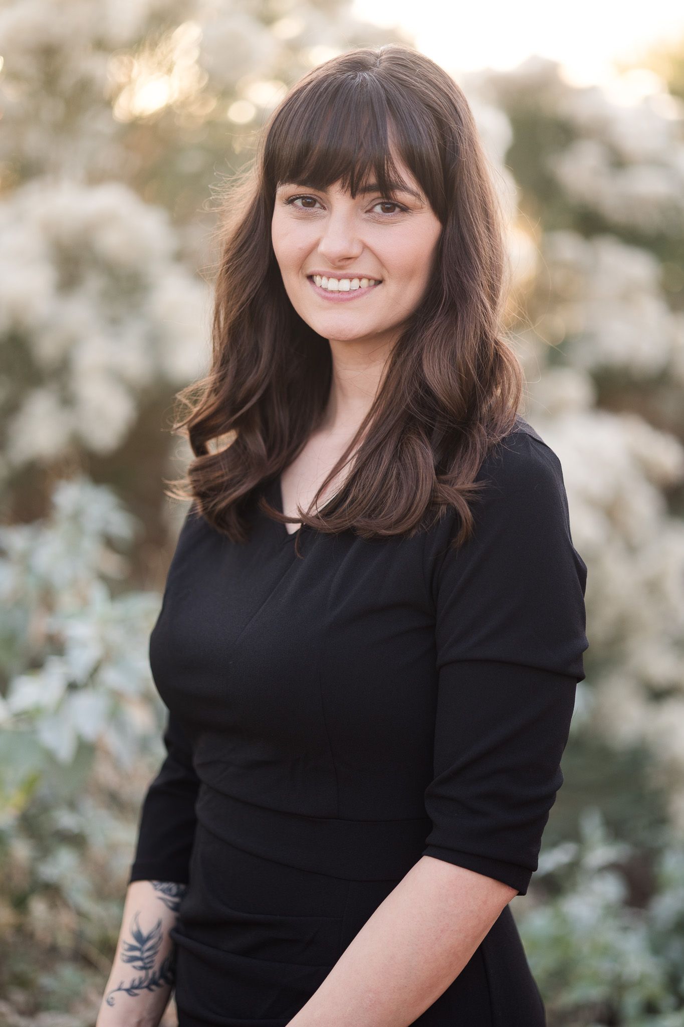 Professional portrait of someone wearing a black top with long dark hair and bangs against a soft blurred natural background.