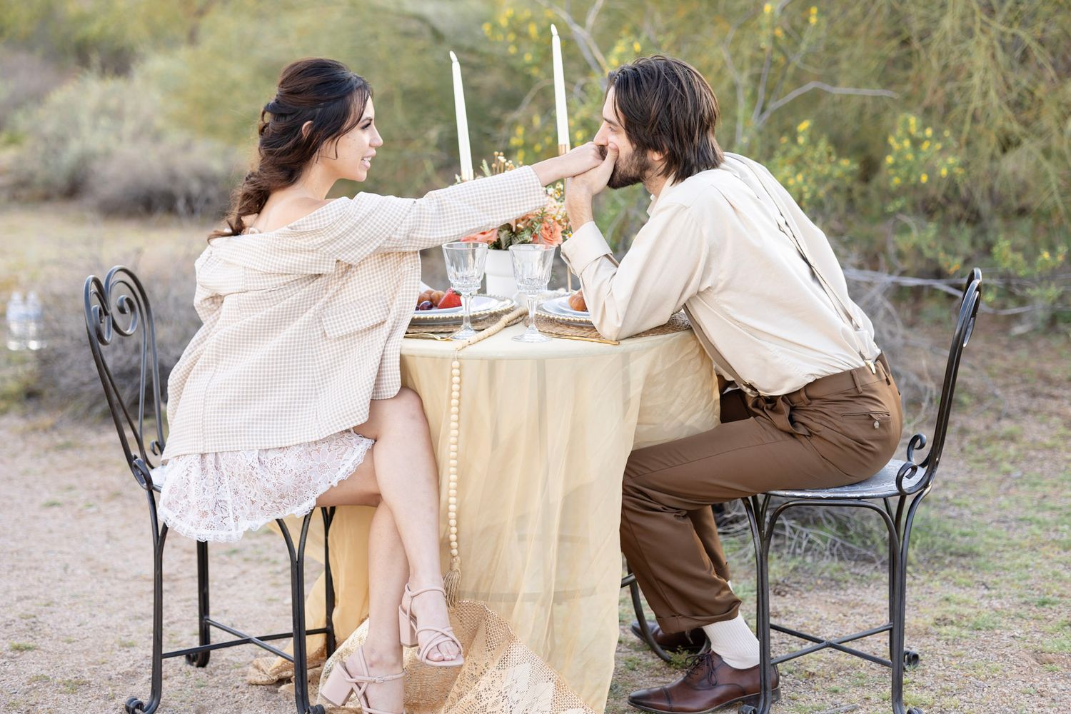 Rustic outdoor dining scene with wrought iron chairs and cream tablecloth in natural setting.