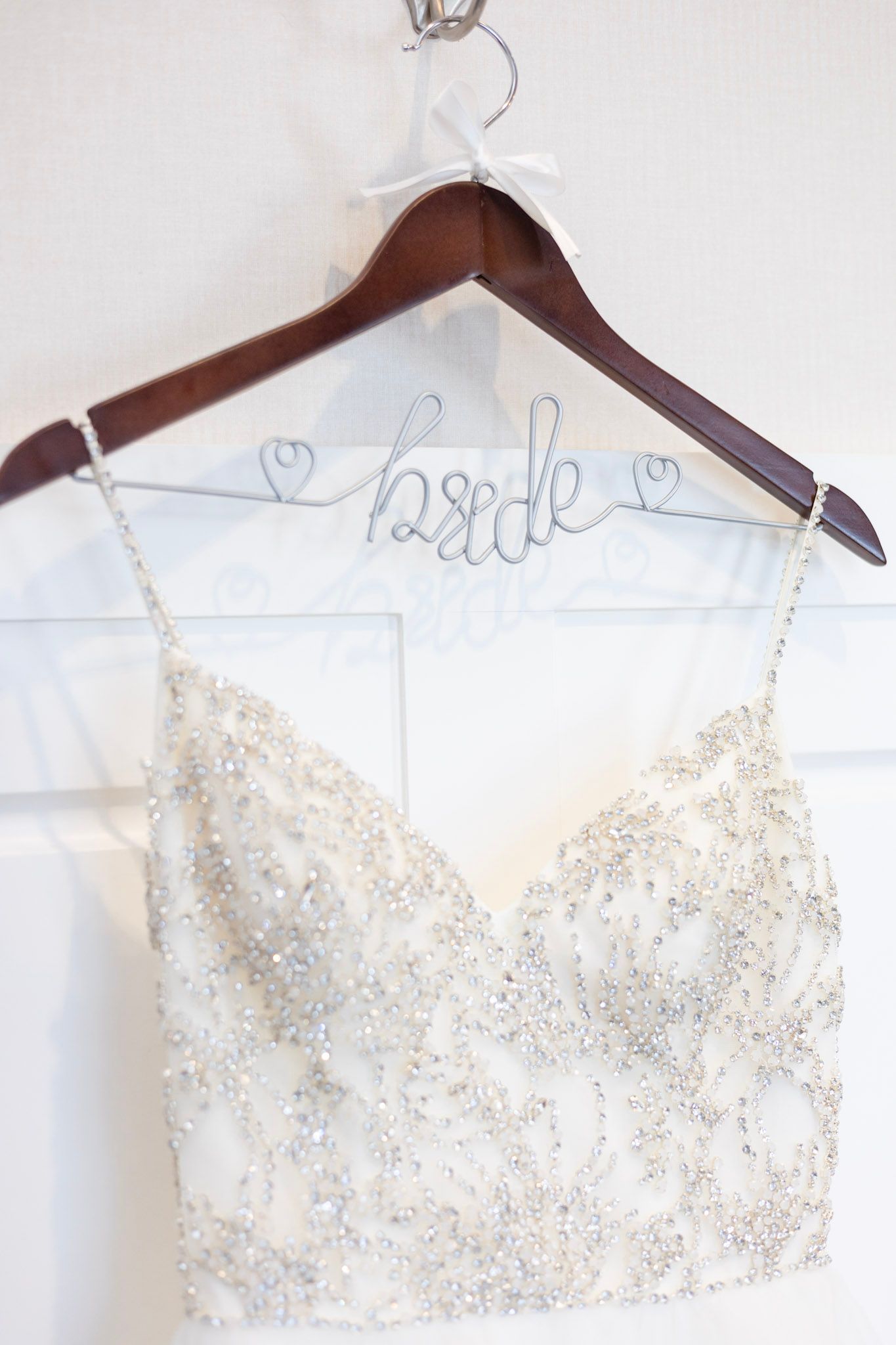 Ornate beaded white wedding dress bodice hanging on wooden hanger against light background.
