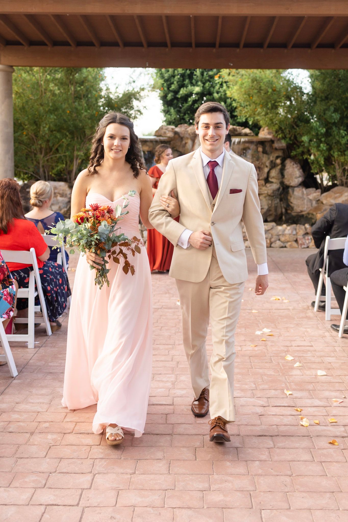 Bridesmaids in blush gowns and groomsmen in beige suits process down a stone patio at an outdoor venue.