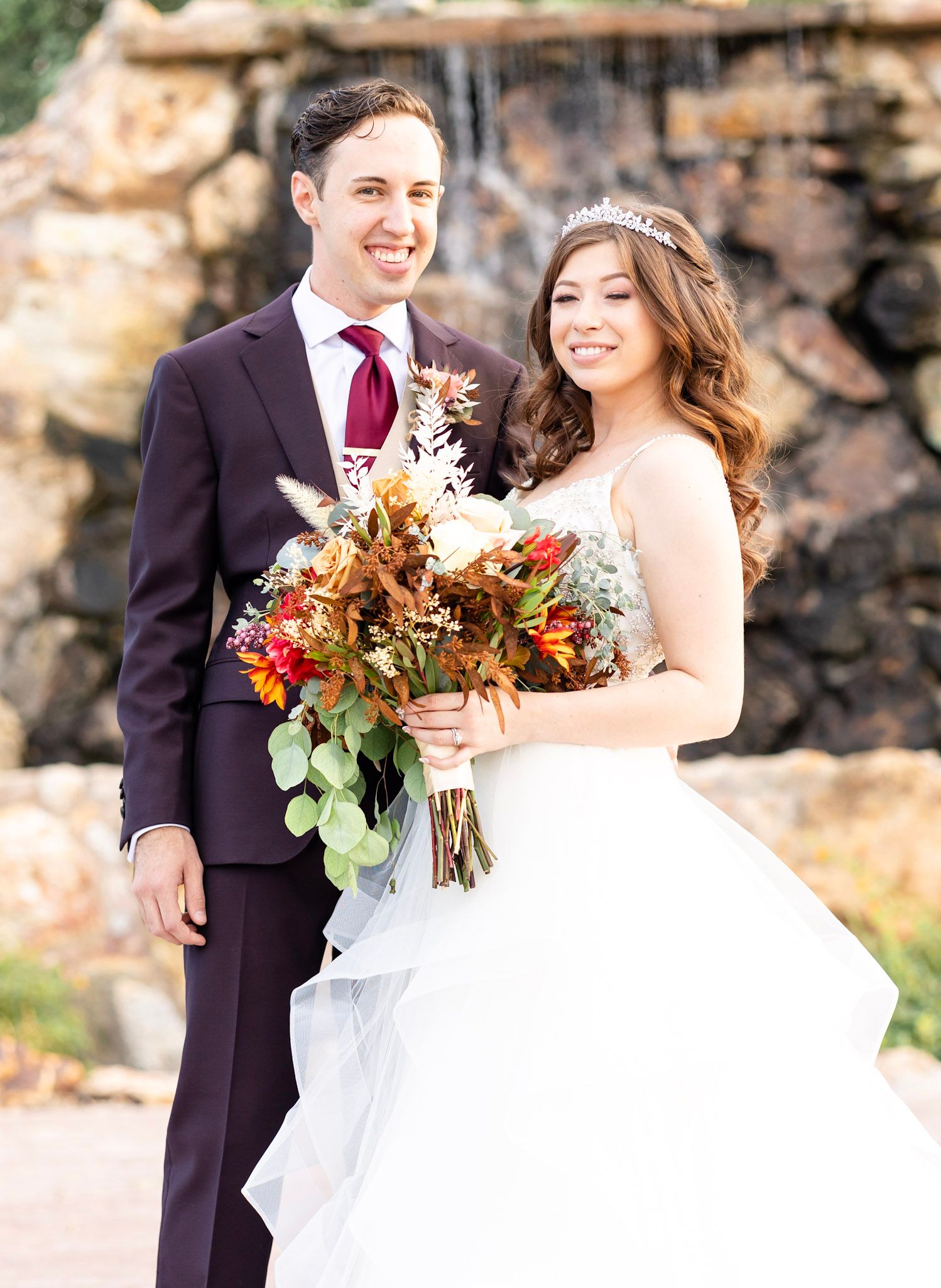 Newlyweds share romantic moment against stone wall with fall-themed floral bouquet.