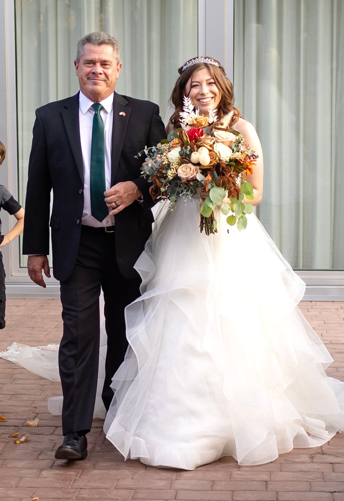 A bride in a flowing white gown walks with her bouquet alongside someone in a dark suit.