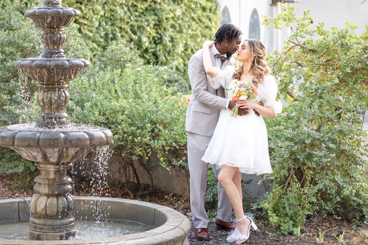 Wedding couple poses near garden fountain in romantic setting.