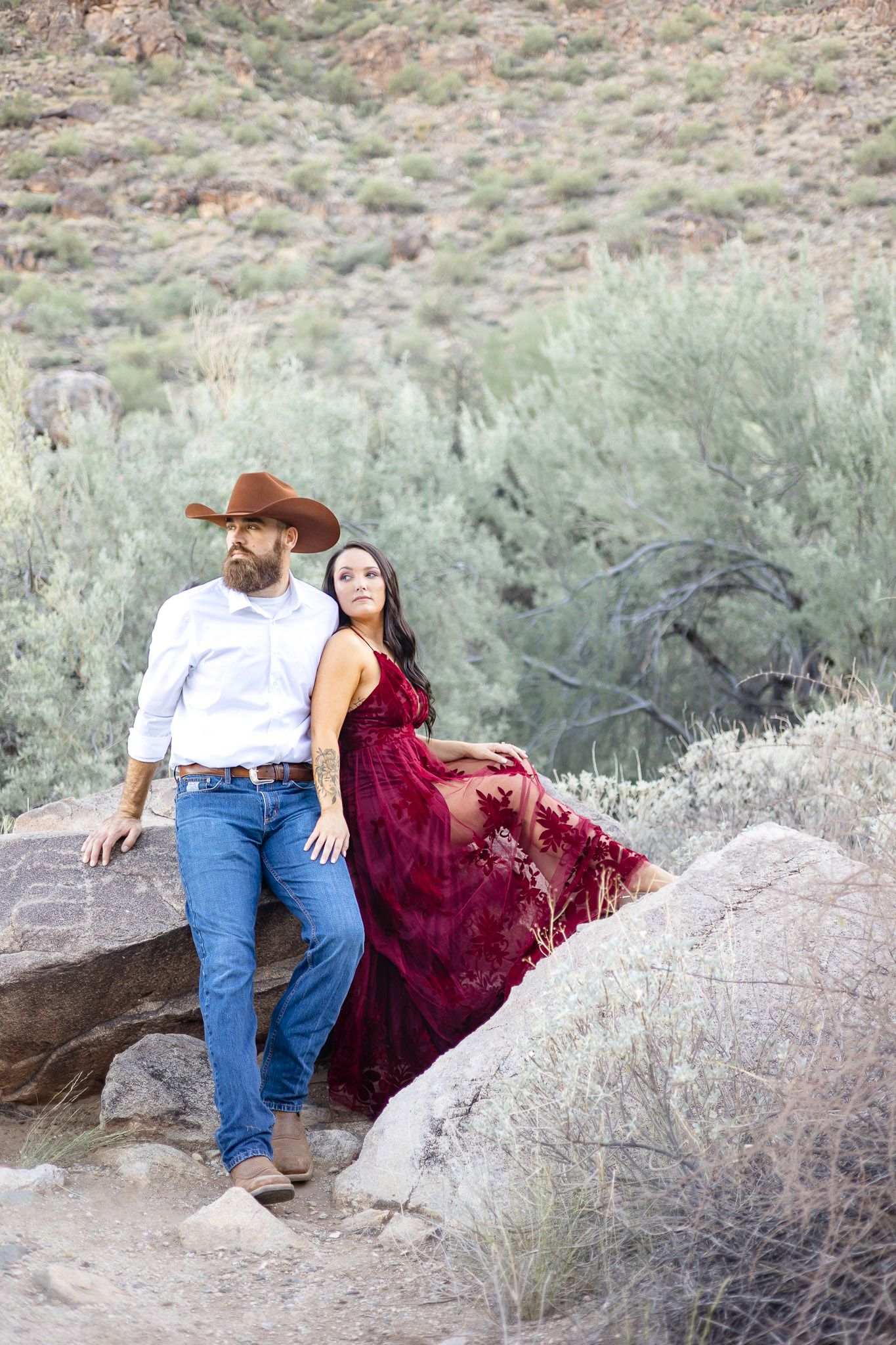 A couple in western attire poses together on desert rocks surrounded by sage brush and rugged terrain.