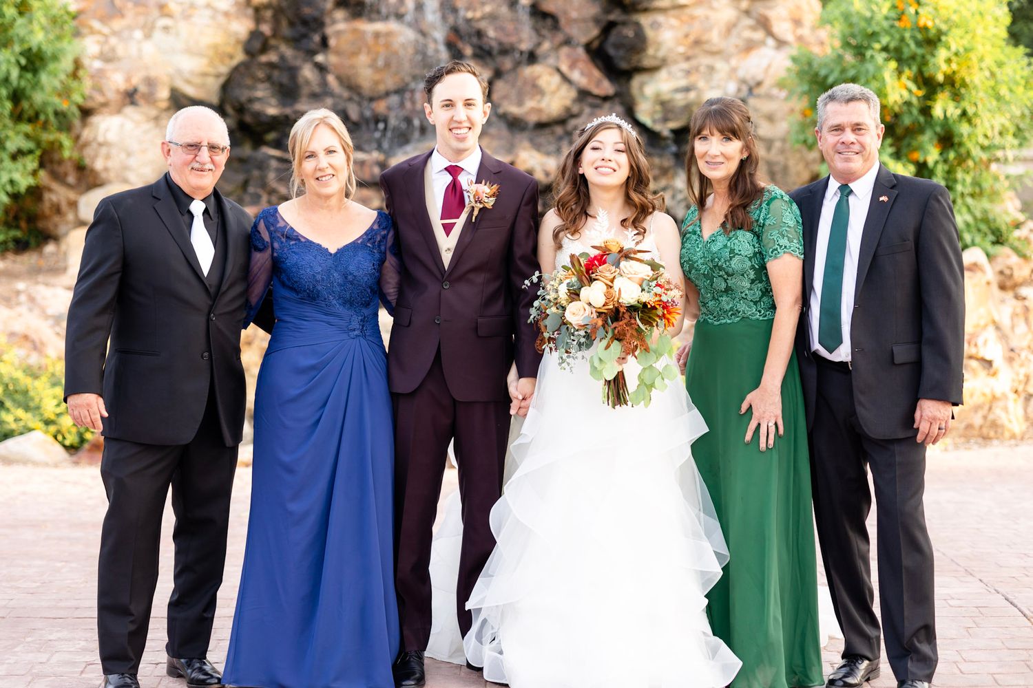 Large wedding party in formal attire poses outdoors with stone wall and greenery.