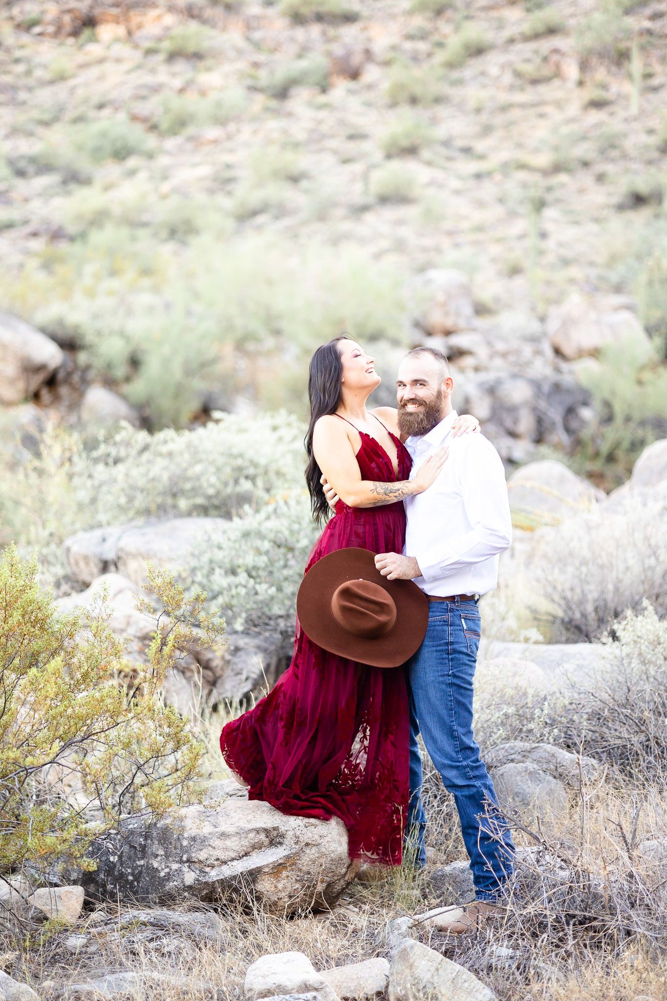 A couple in desert terrain share romantic moments as she wears a burgundy dress and he wears jeans and a white shirt.