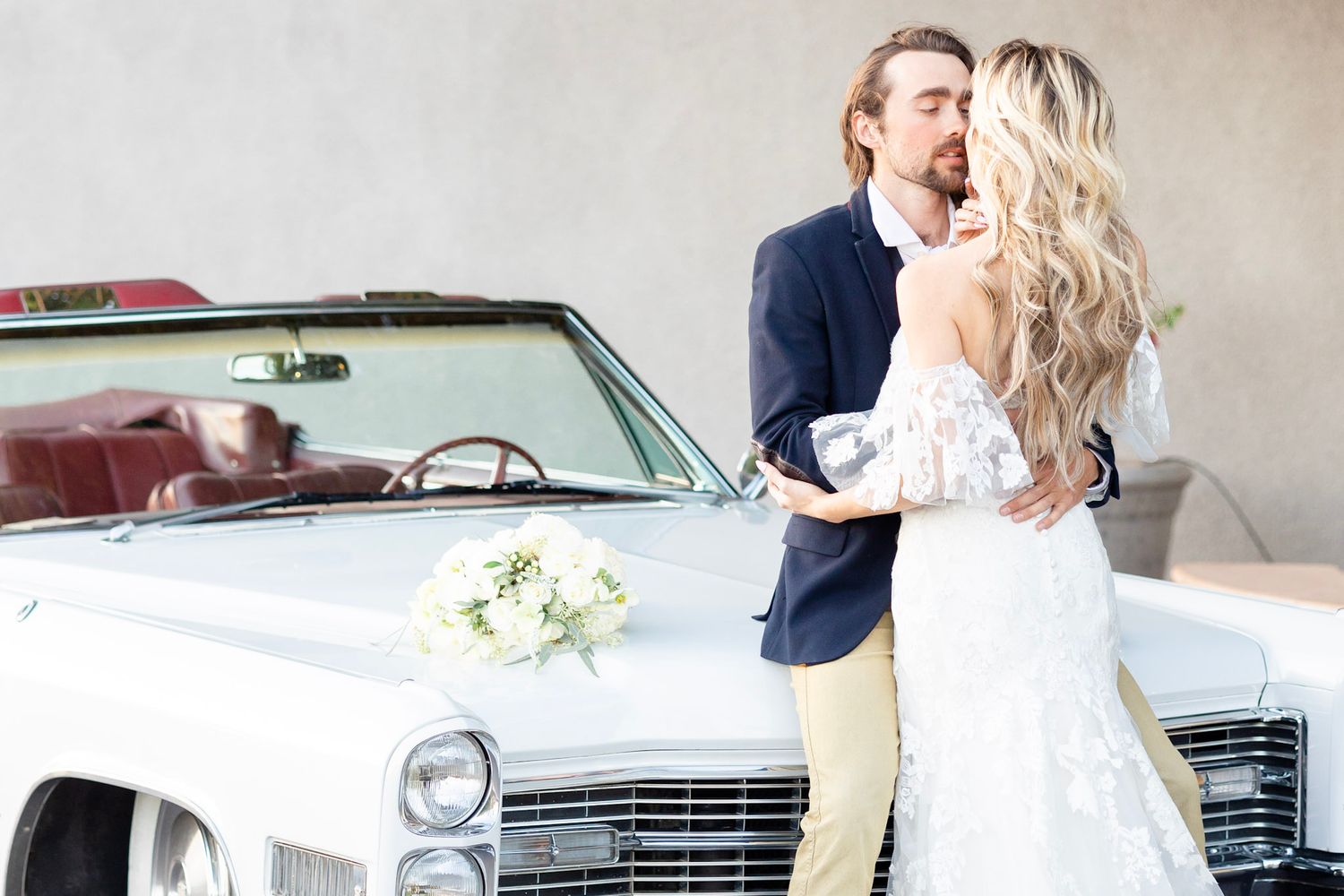 Elegant wedding couple poses next to vintage white car with floral decorations.