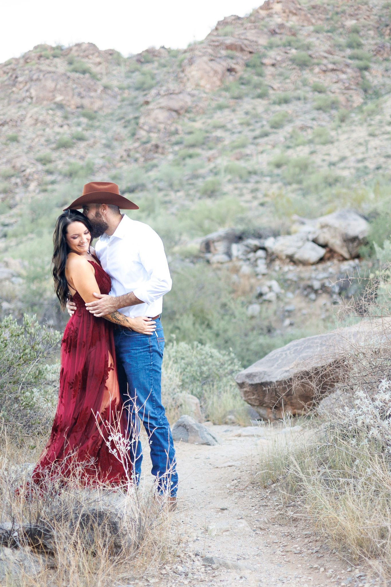 A couple shares romantic moments along a desert trail with mountain views in the background.