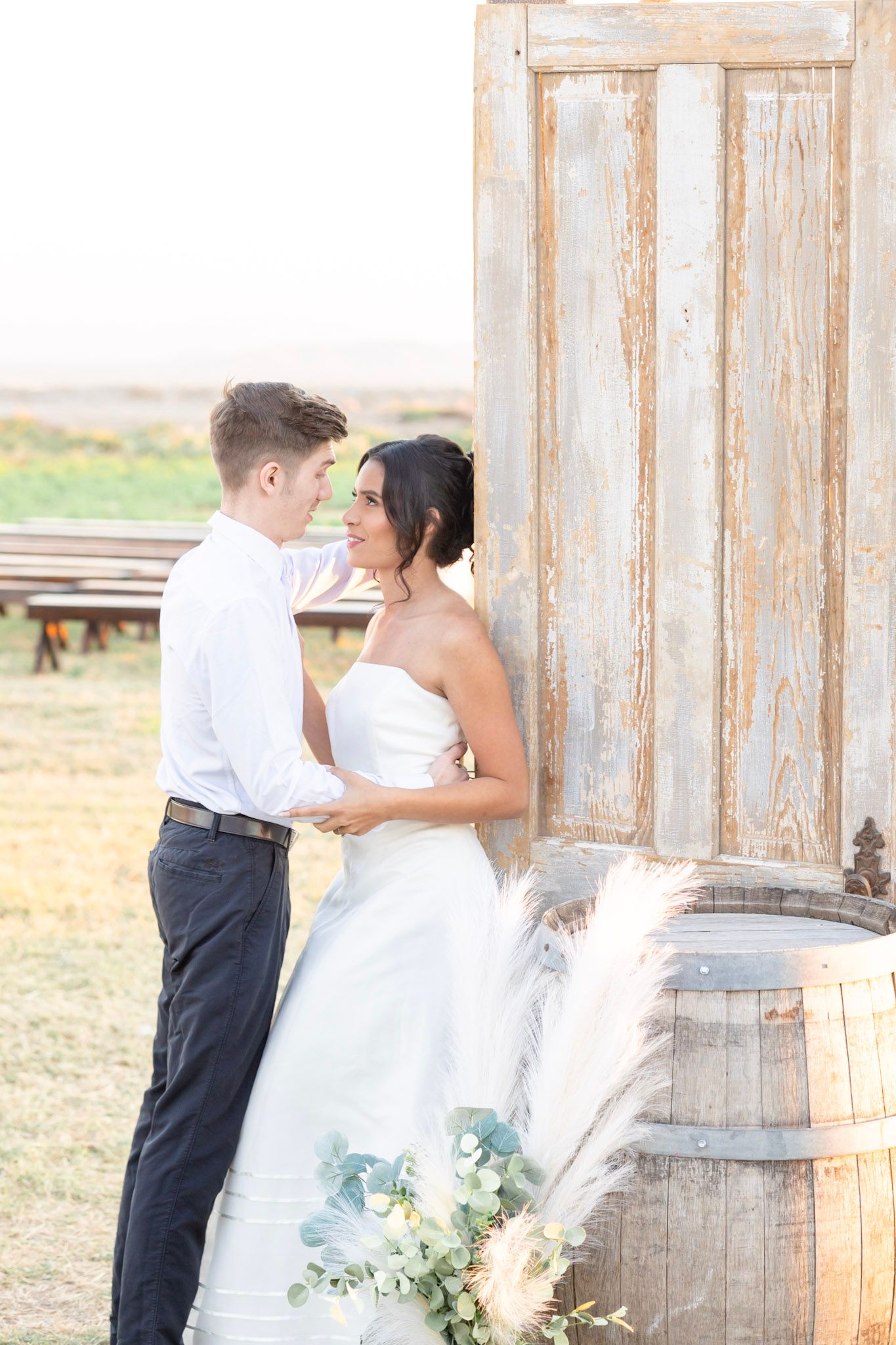 Romantic wedding photoshoot near rustic wooden walls and vintage barrels in a countryside setting.
