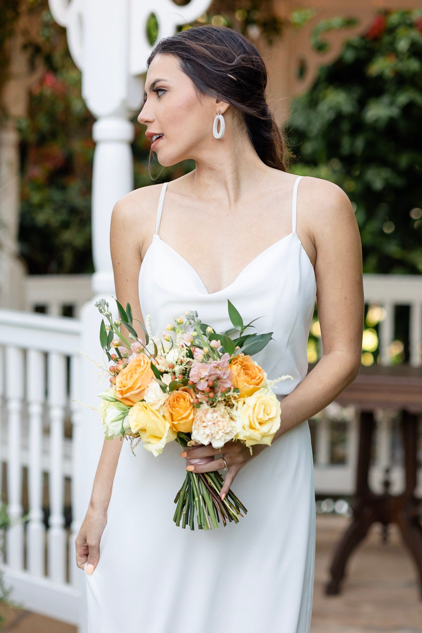 Bride in white dress holding peach and yellow rose bouquet against garden background.