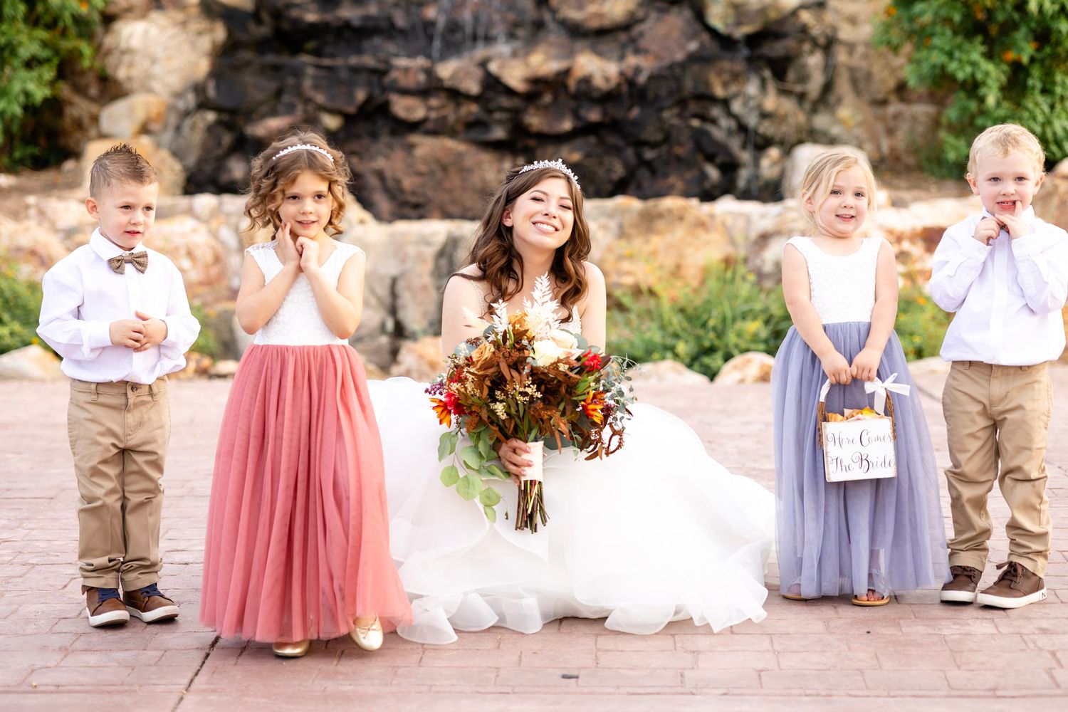 Wedding party with children in pastel dresses poses on stone patio during outdoor ceremony.