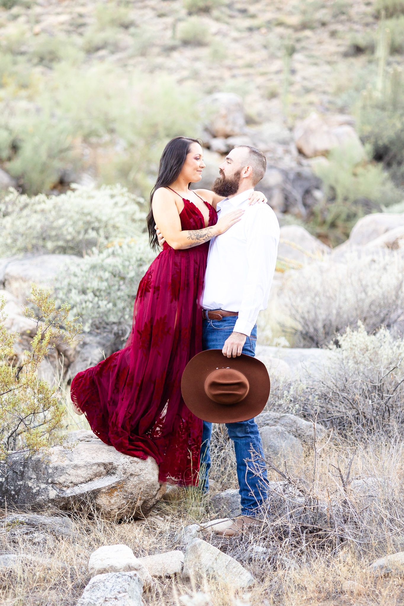 A couple in a burgundy dress and cowboy attire share romantic moments among desert brush in a rustic outdoor setting.