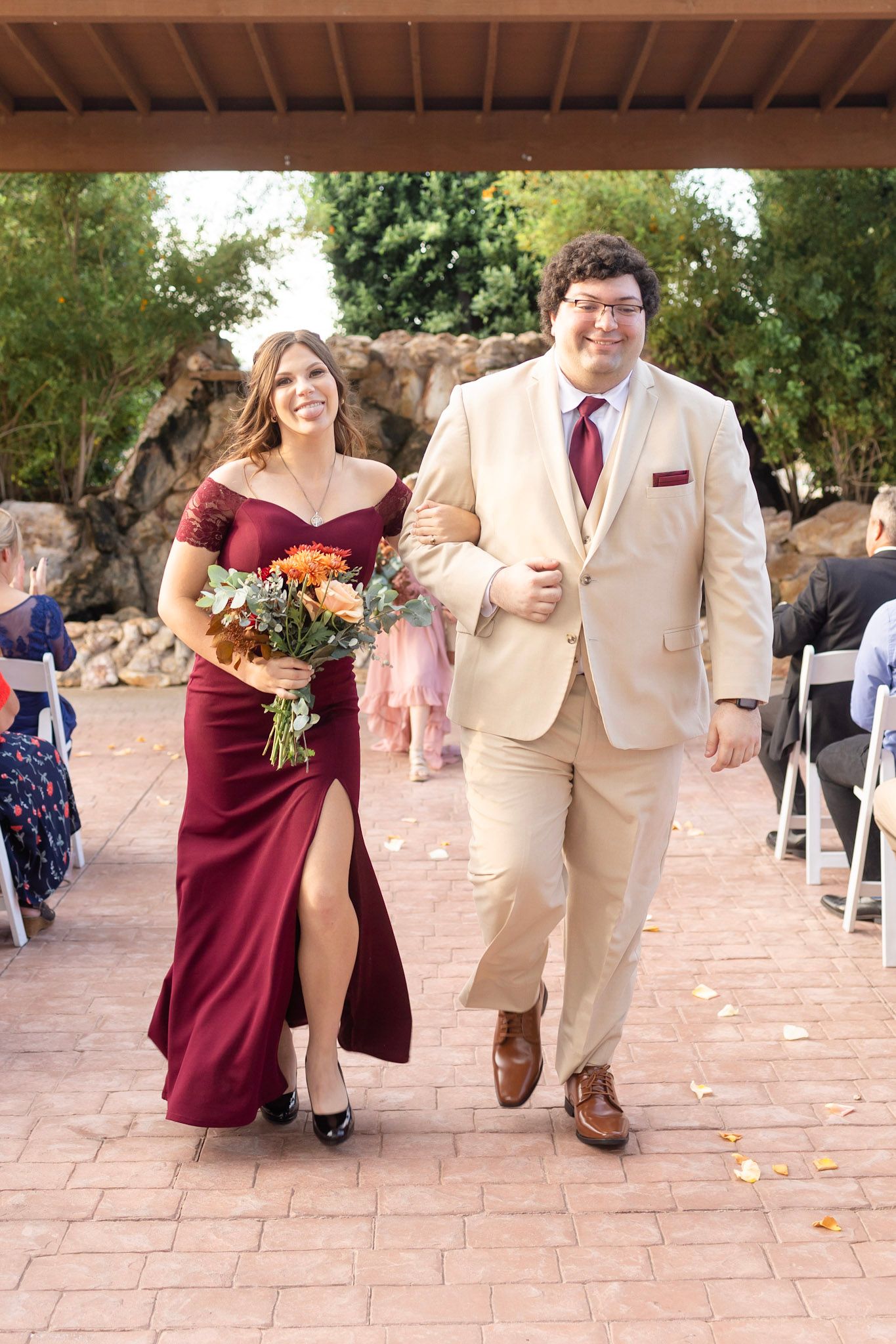 Wedding party members in maroon dresses and khaki suits process down an outdoor brick pathway.