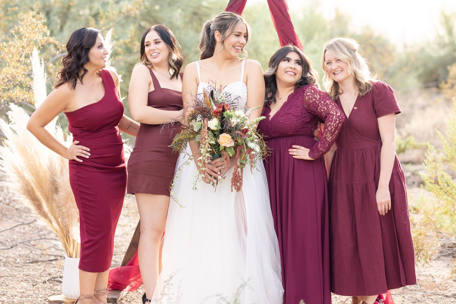 Bridesmaids in burgundy dresses gather around bride for outdoor wedding photos.
