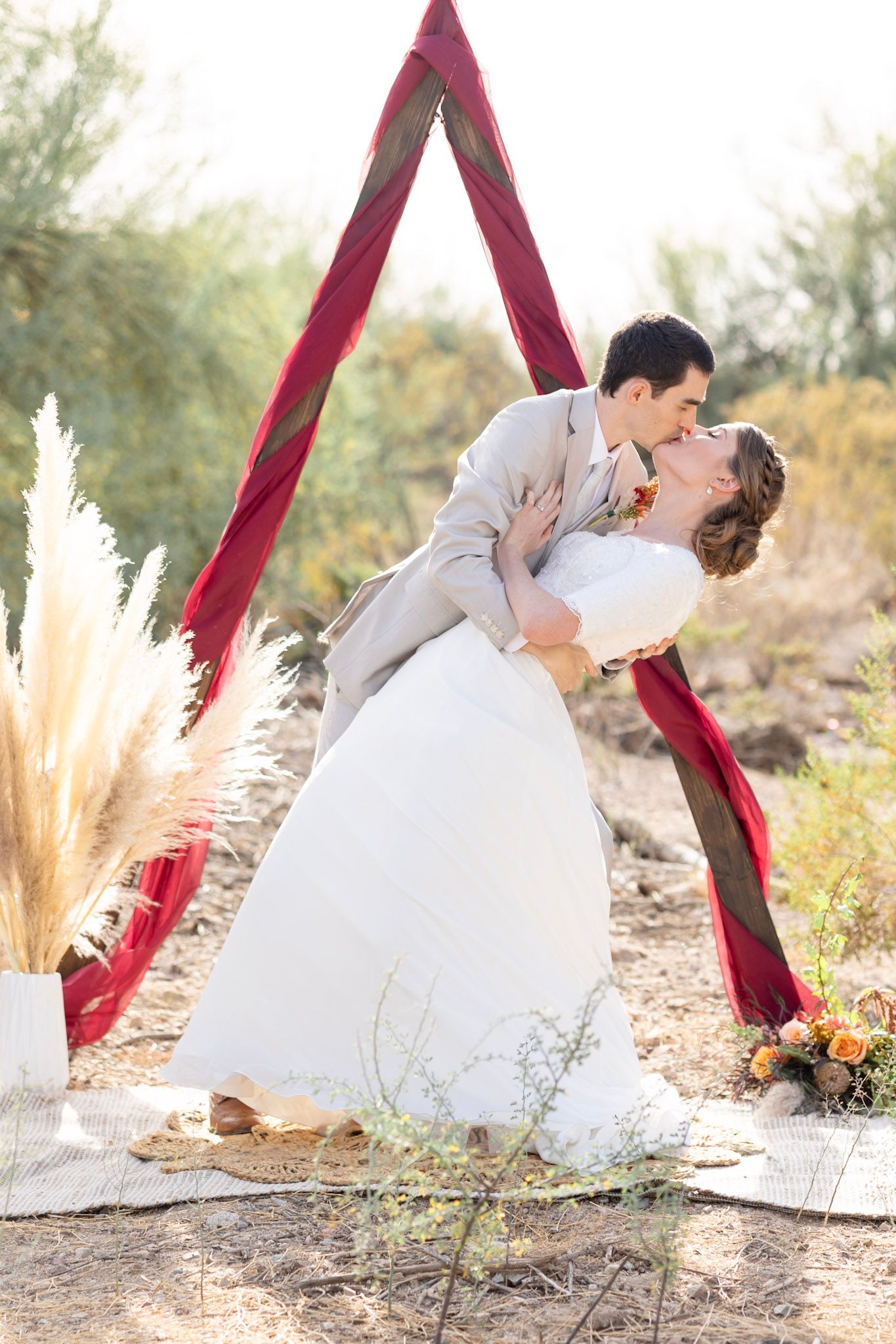 Romantic wedding couple dips and kisses under red fabric draping in desert setting with pampas grass decor.