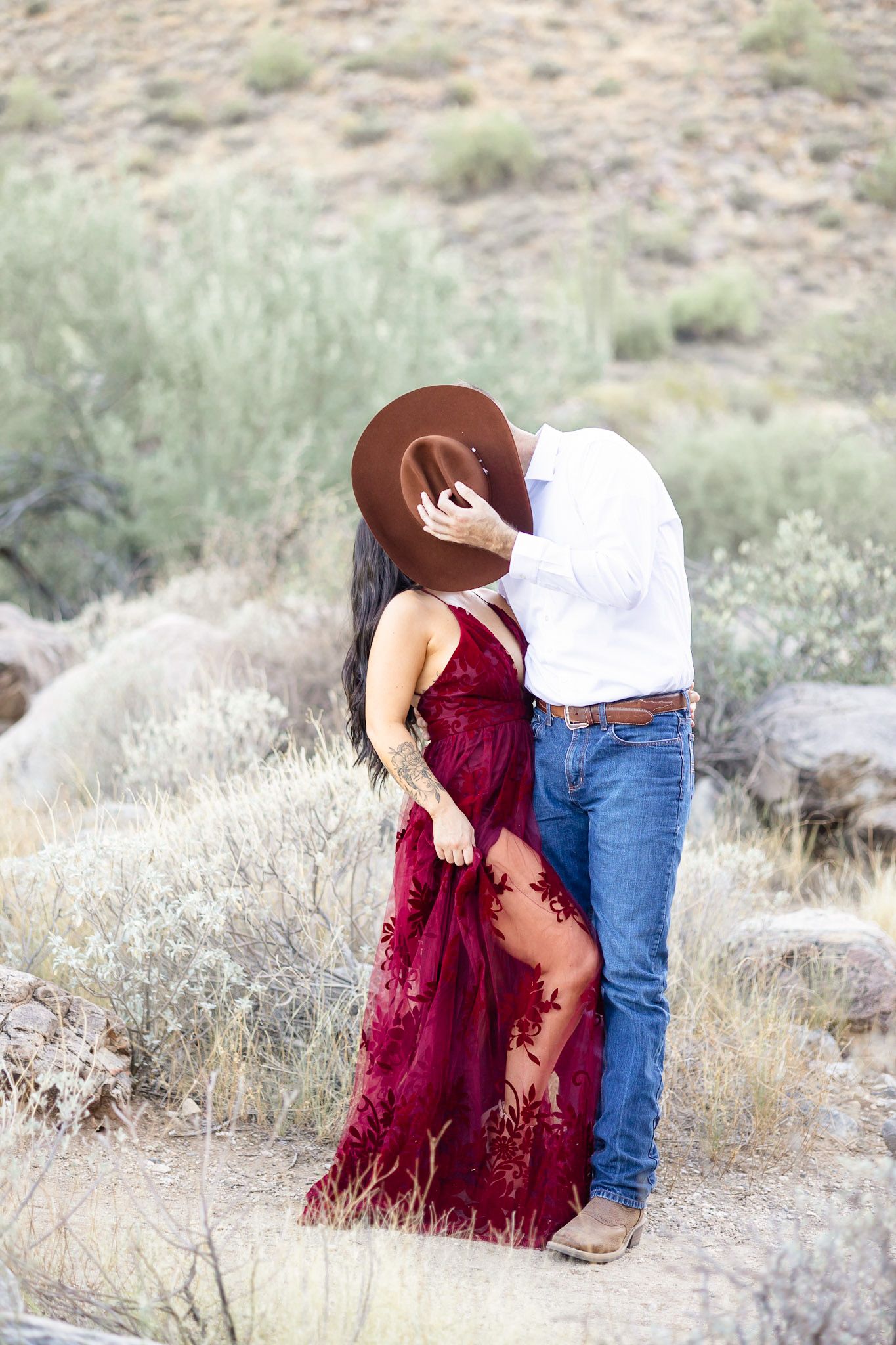 A couple in a burgundy dress and jeans embrace in a desert landscape with desert brush and scrub vegetation.