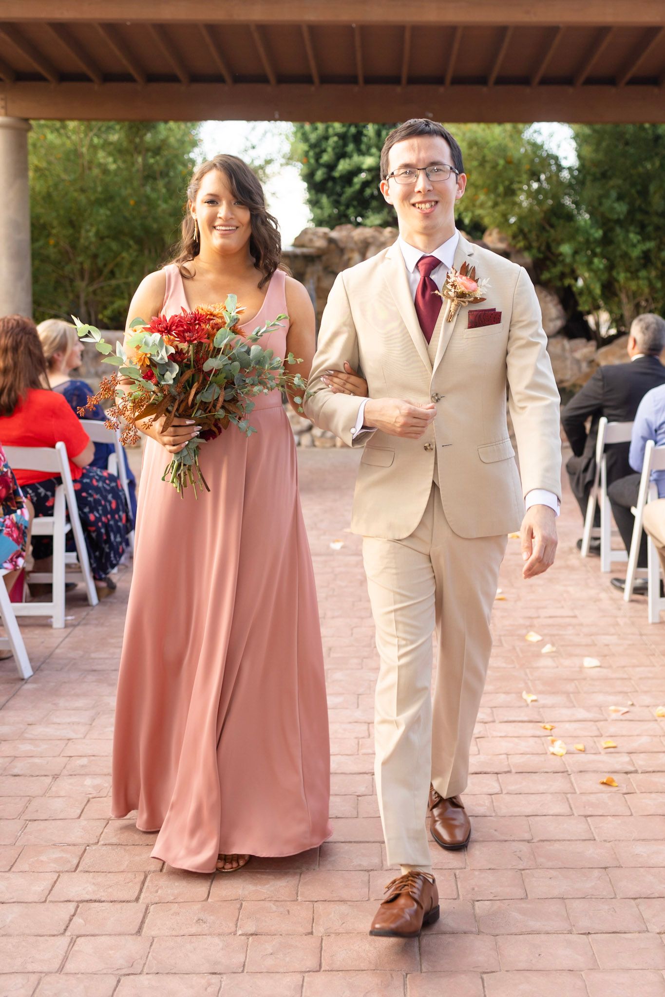 Wedding party members in pink dresses and tan suits walk down a brick pathway during an outdoor ceremony.