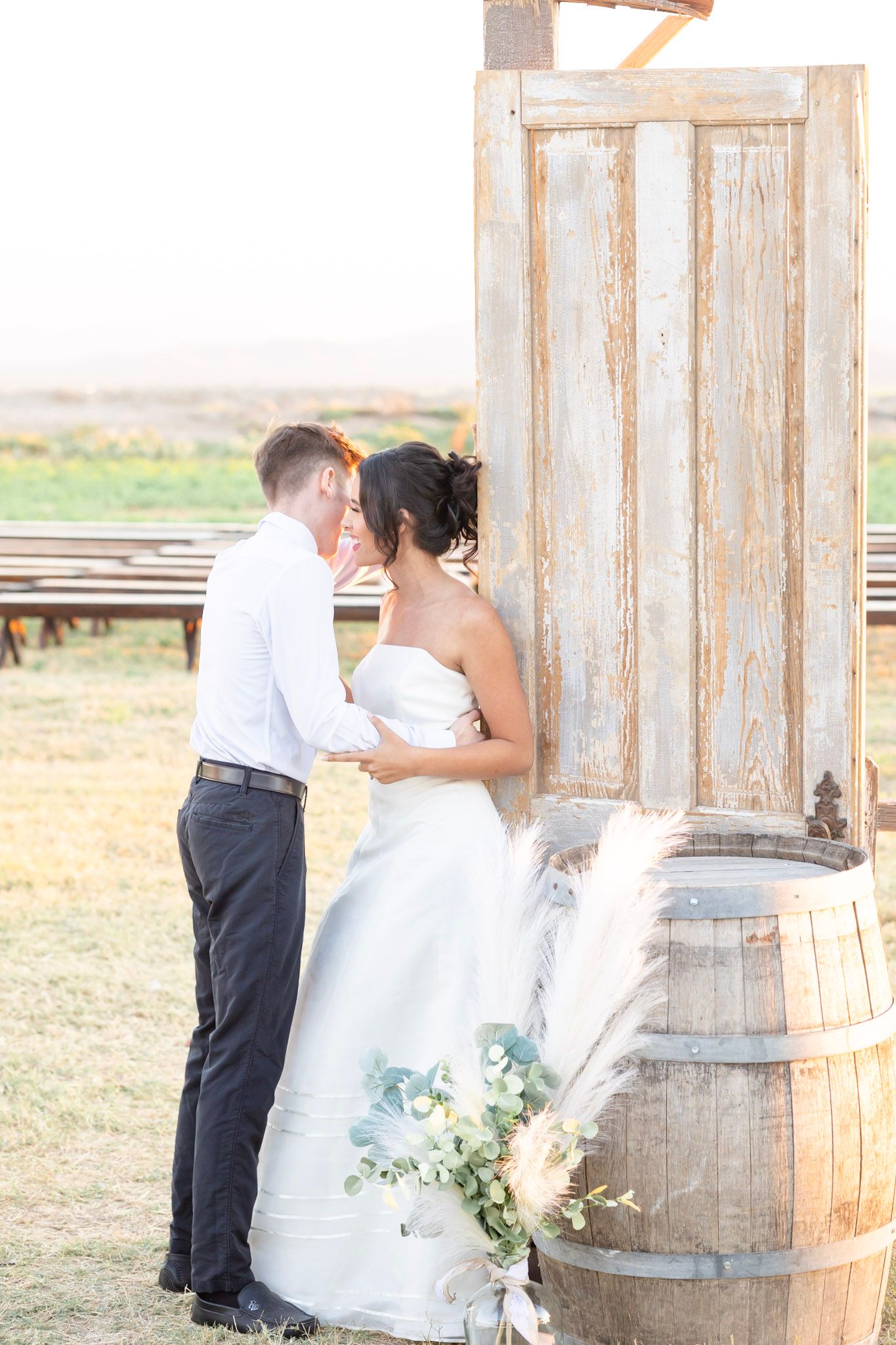 Wedding couple embraces against weathered wooden backdrop with rustic barrels in natural light.