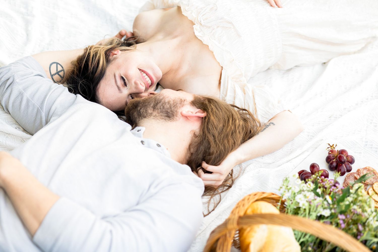 A person with long blonde hair lies peacefully in white bedding surrounded by fresh fruits and flowers.