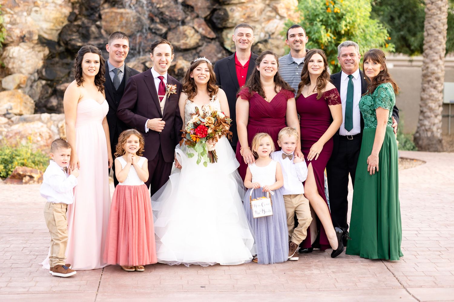 Wedding group photo with bridesmaids in pink and green dresses against stone wall.