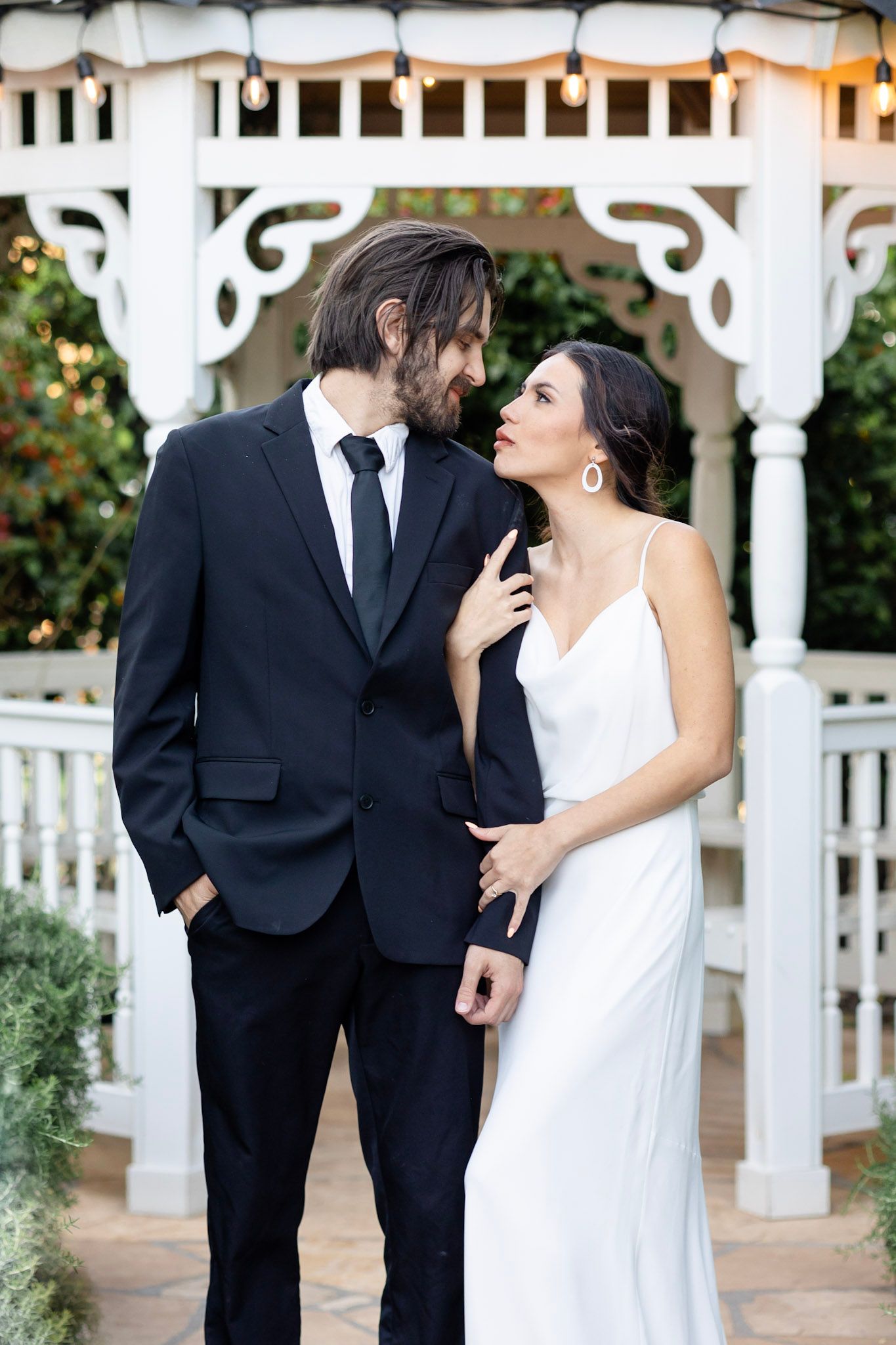 Romantic wedding couple poses by white gazebo in formal attire.