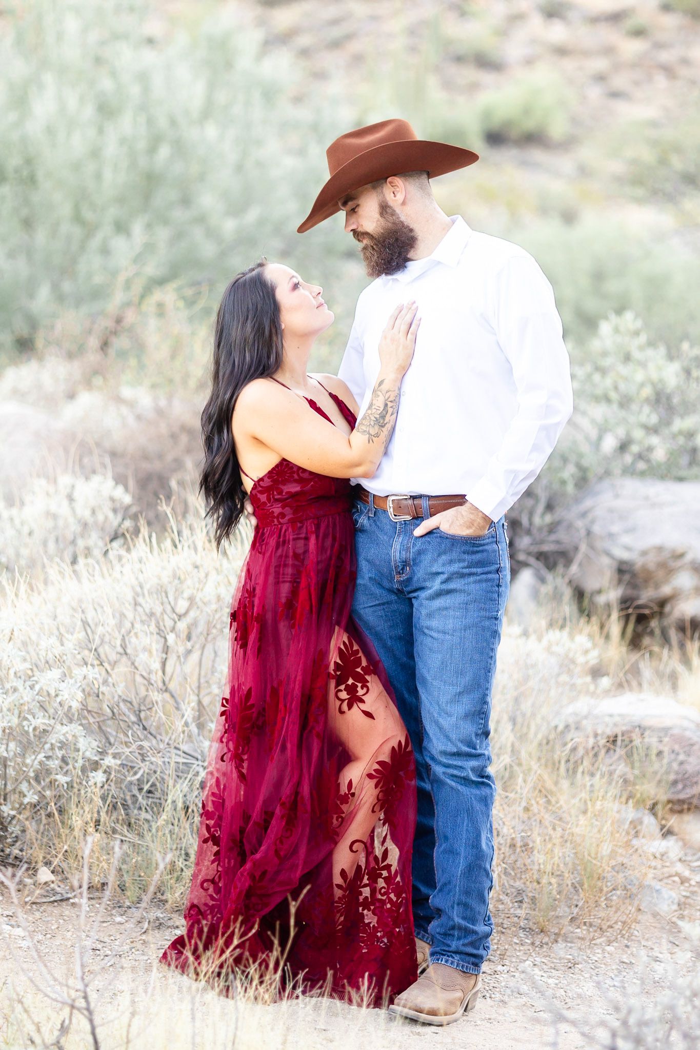 A romantic couple embraces in a desert landscape wearing burgundy dress and cowboy attire at sunset.