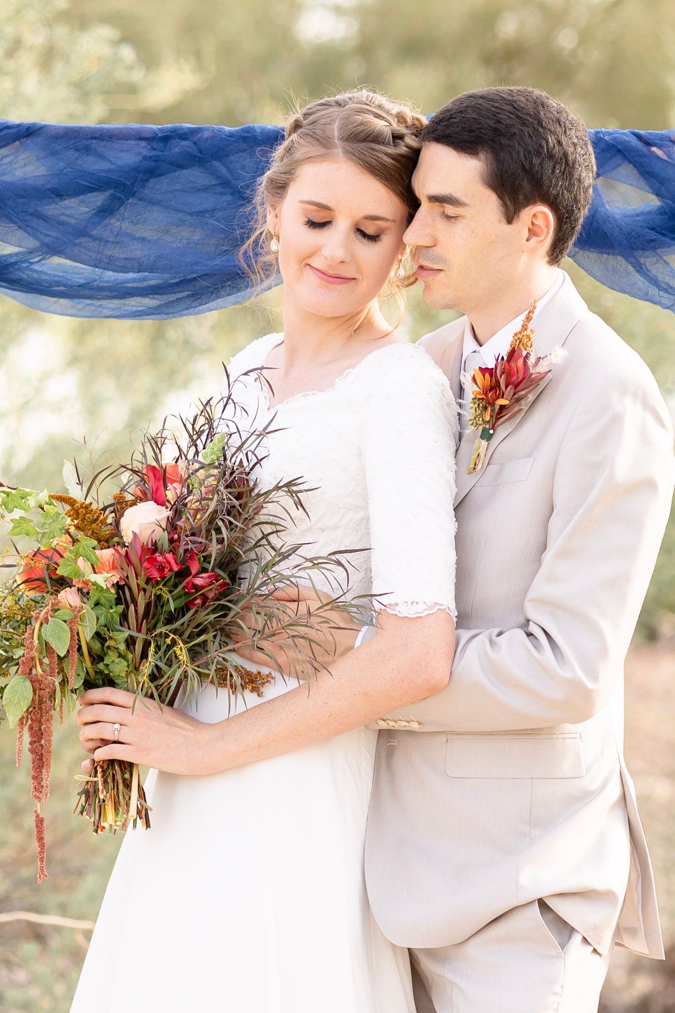 Bride in white dress holds autumn-colored bouquet while embracing groom in light grey suit.